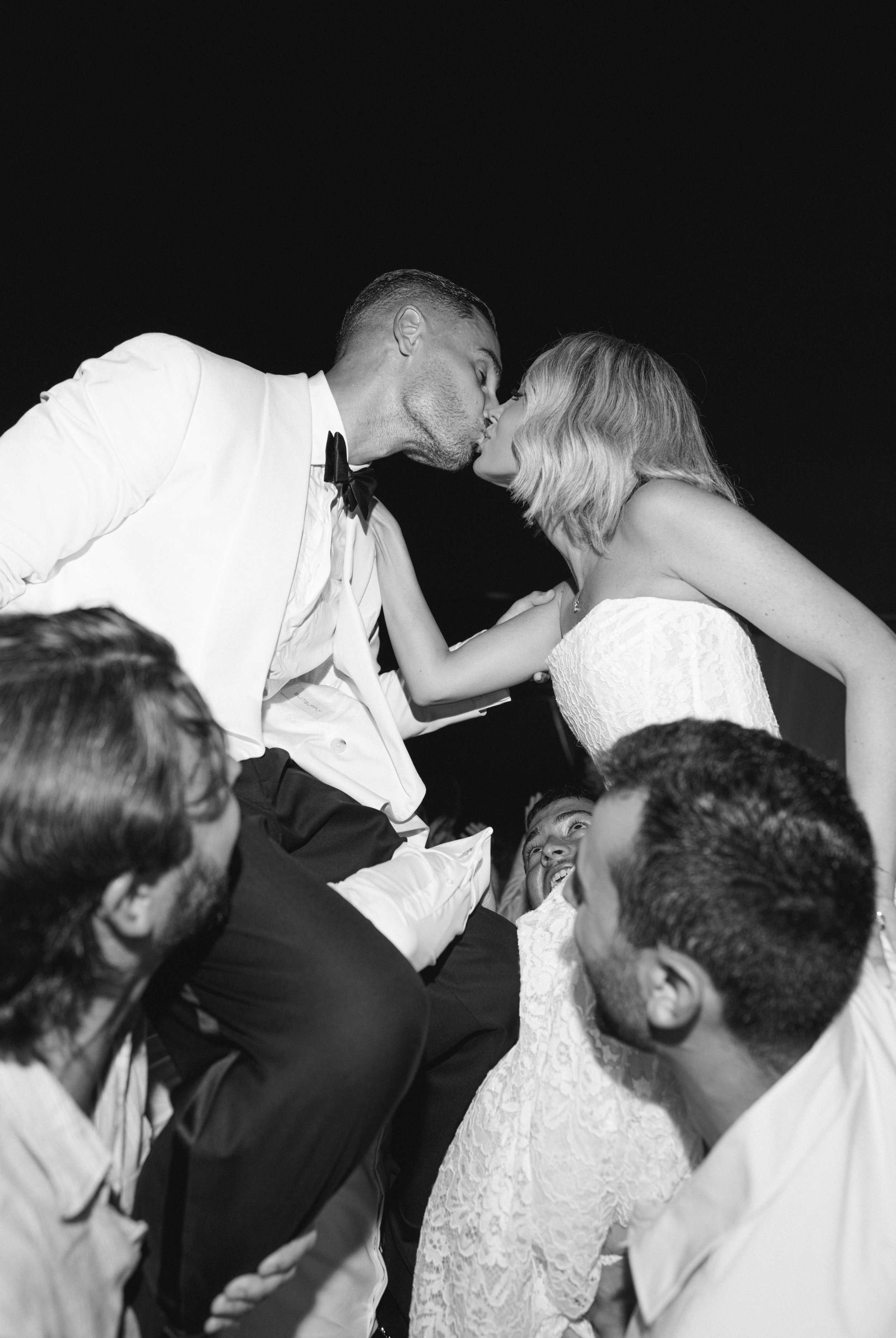 A couple in wedding attire sharing a kiss at night, surrounded by friends at a celebration.