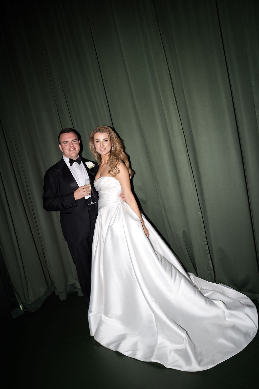A bride and groom standing together at a wedding reception, dressed in formal wedding attire, with the groom holding a champagne glass.