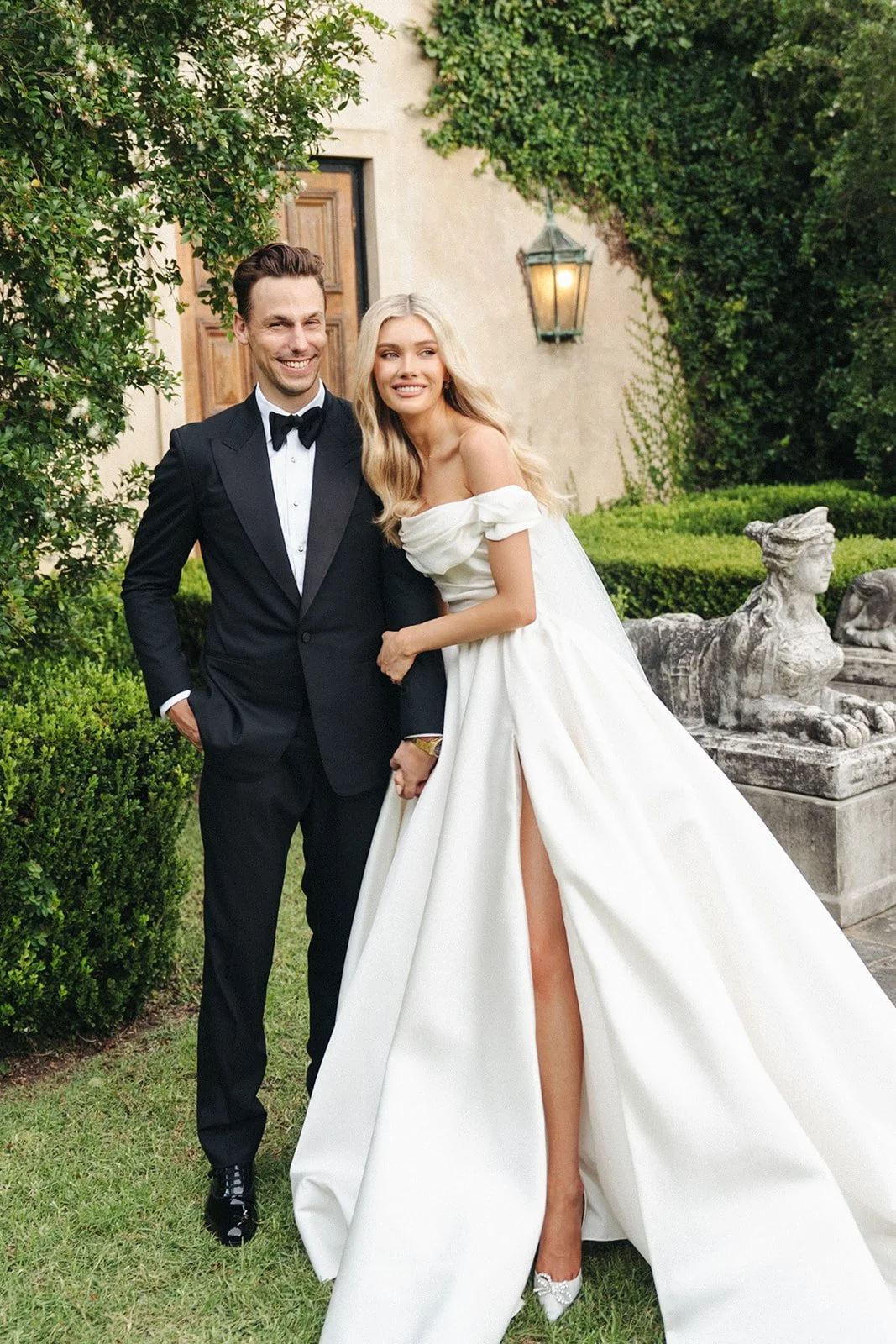 A smiling bride and groom standing together in a garden beside an ivy-covered building.