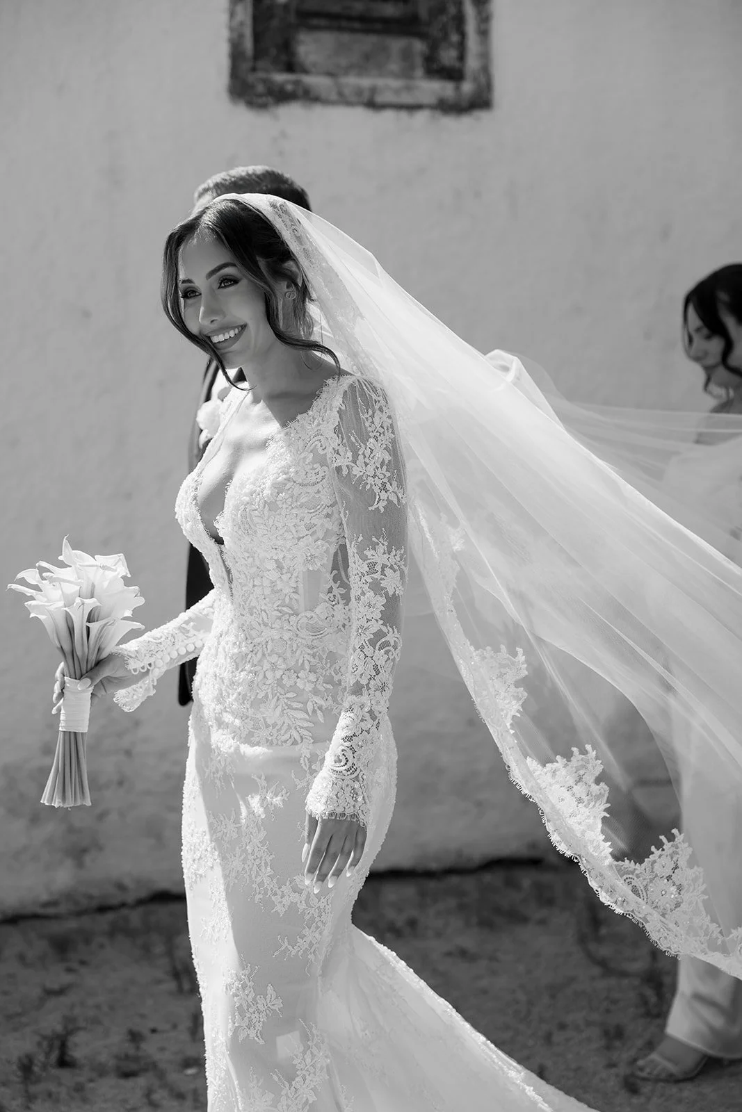 Black and white photo of a bride in a lace wedding dress holding a bouquet of calla lilies, smiling, with a long veil flowing behind her, standing outside.