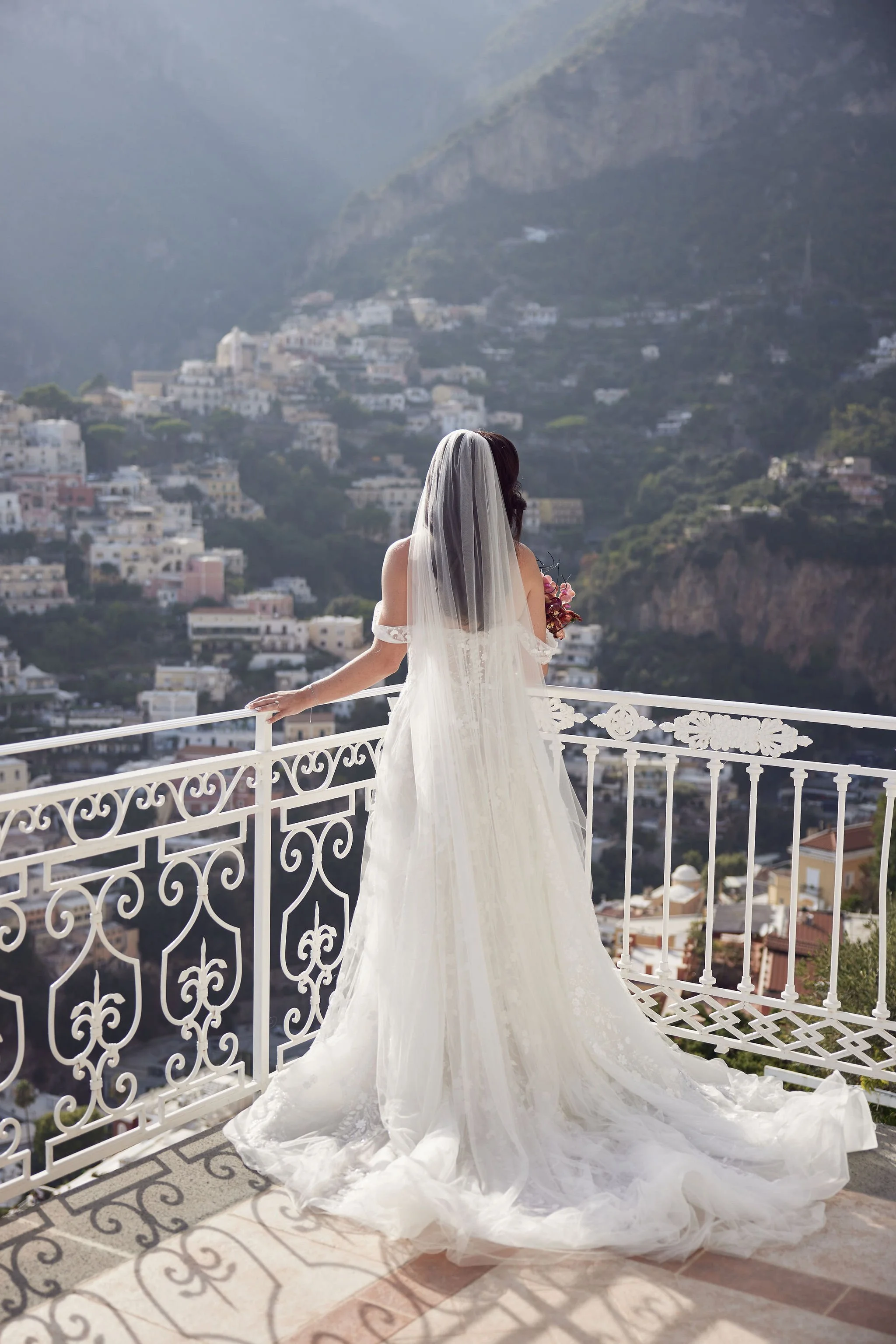 Bride with a long train and veil standing on a balcony overlooking a hillside town and mountains.