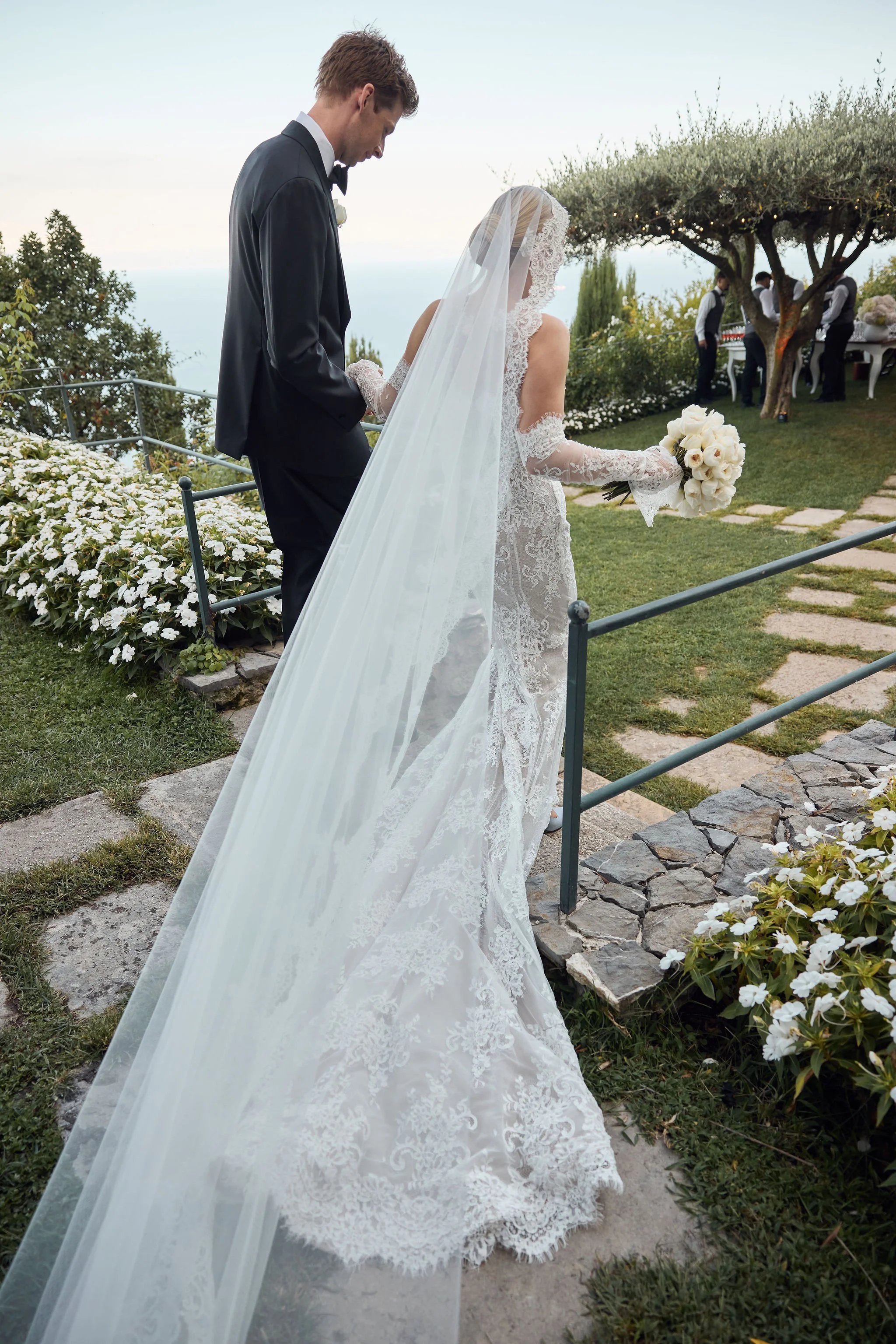 A bride in a lace wedding dress and veil holding a bouquet of white flowers, walking with a groom in a black tuxedo during an outdoor wedding ceremony.