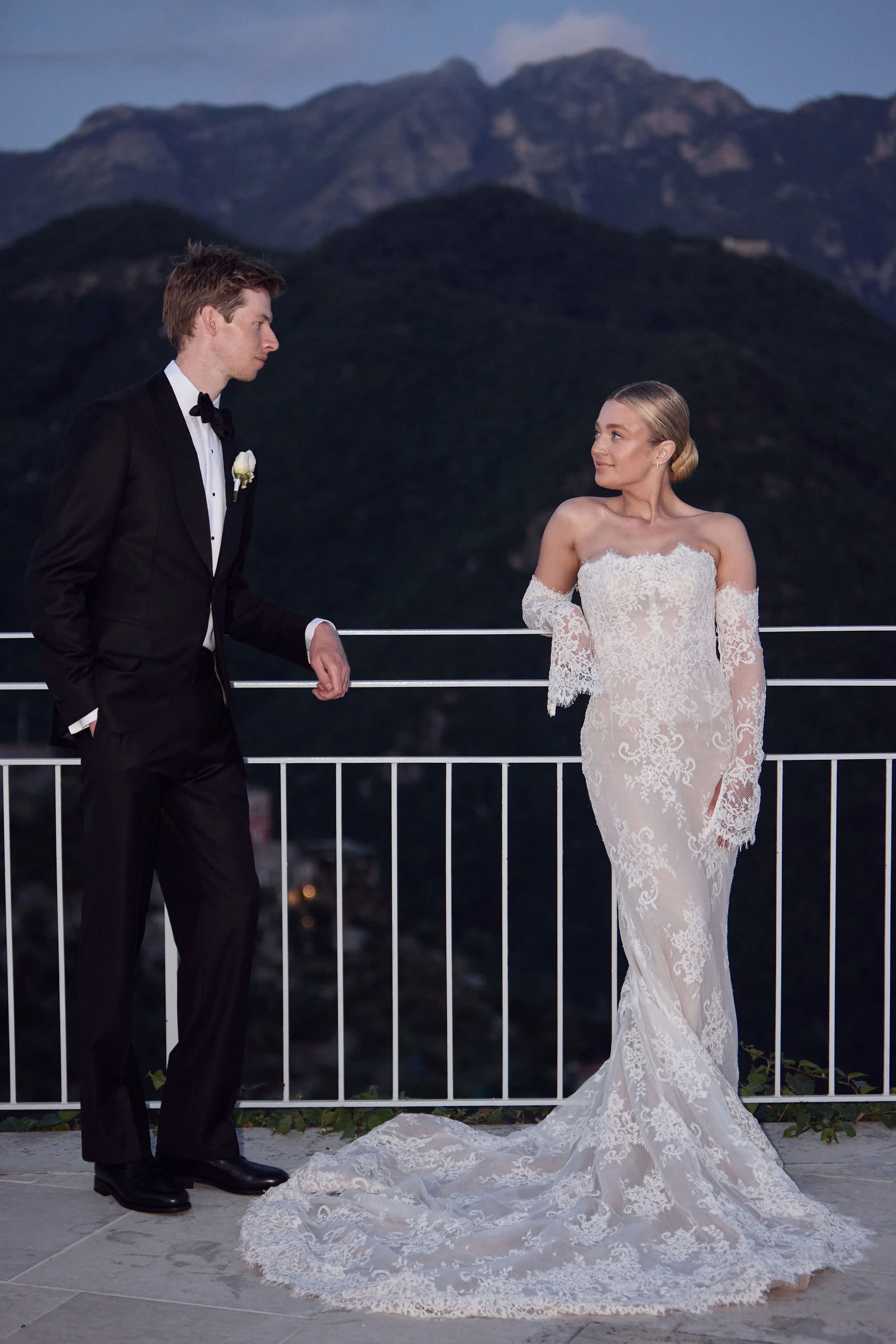 A bride and groom standing on a balcony at sunset, overlooking mountains. The groom is in a black tuxedo, and the bride is in a lace wedding dress with off-the-shoulder sleeves.