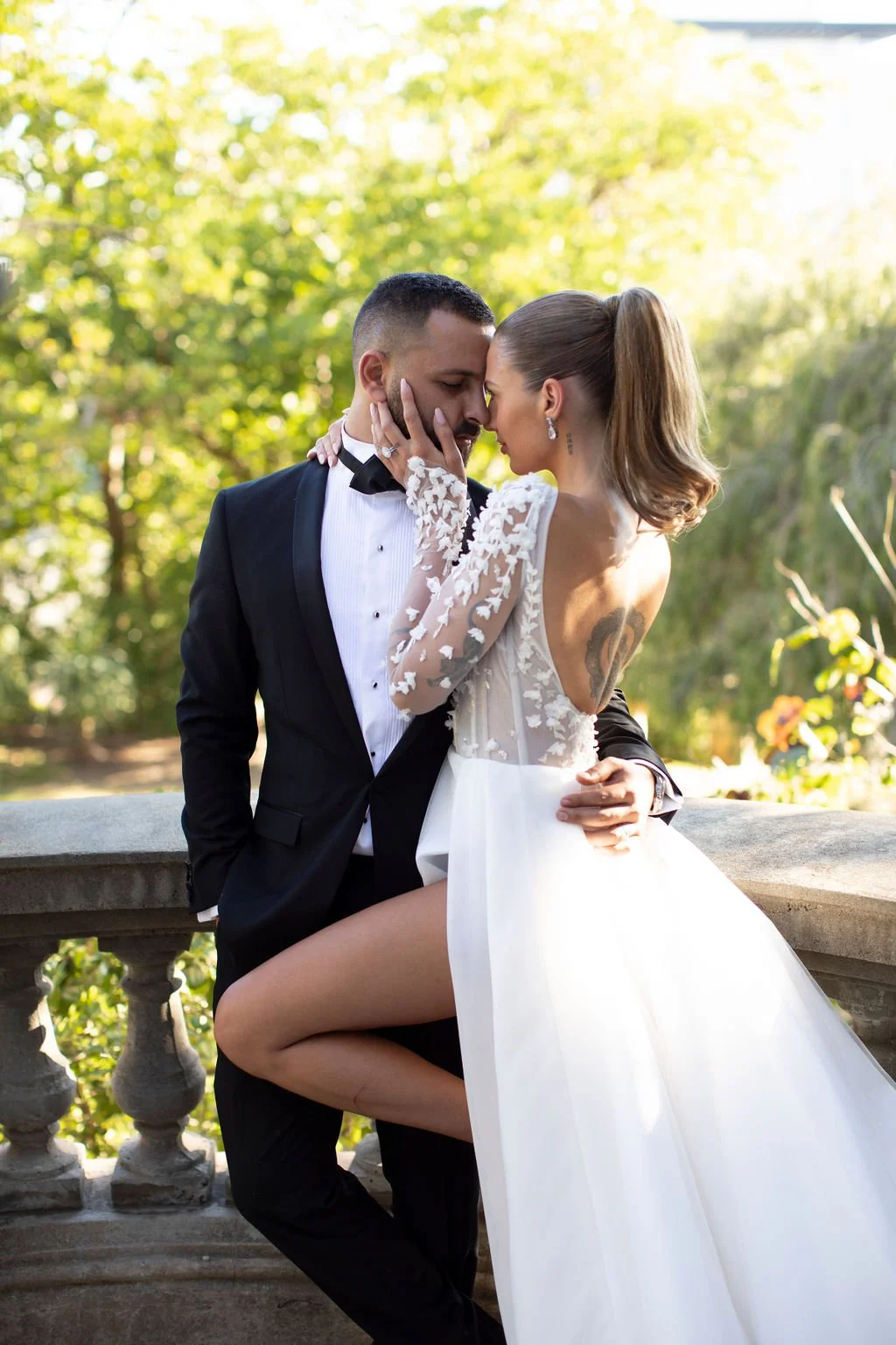 A bride and groom in wedding attire embrace outdoors on a sunny day, with the bride's knee resting on the groom's thigh and their foreheads touching.