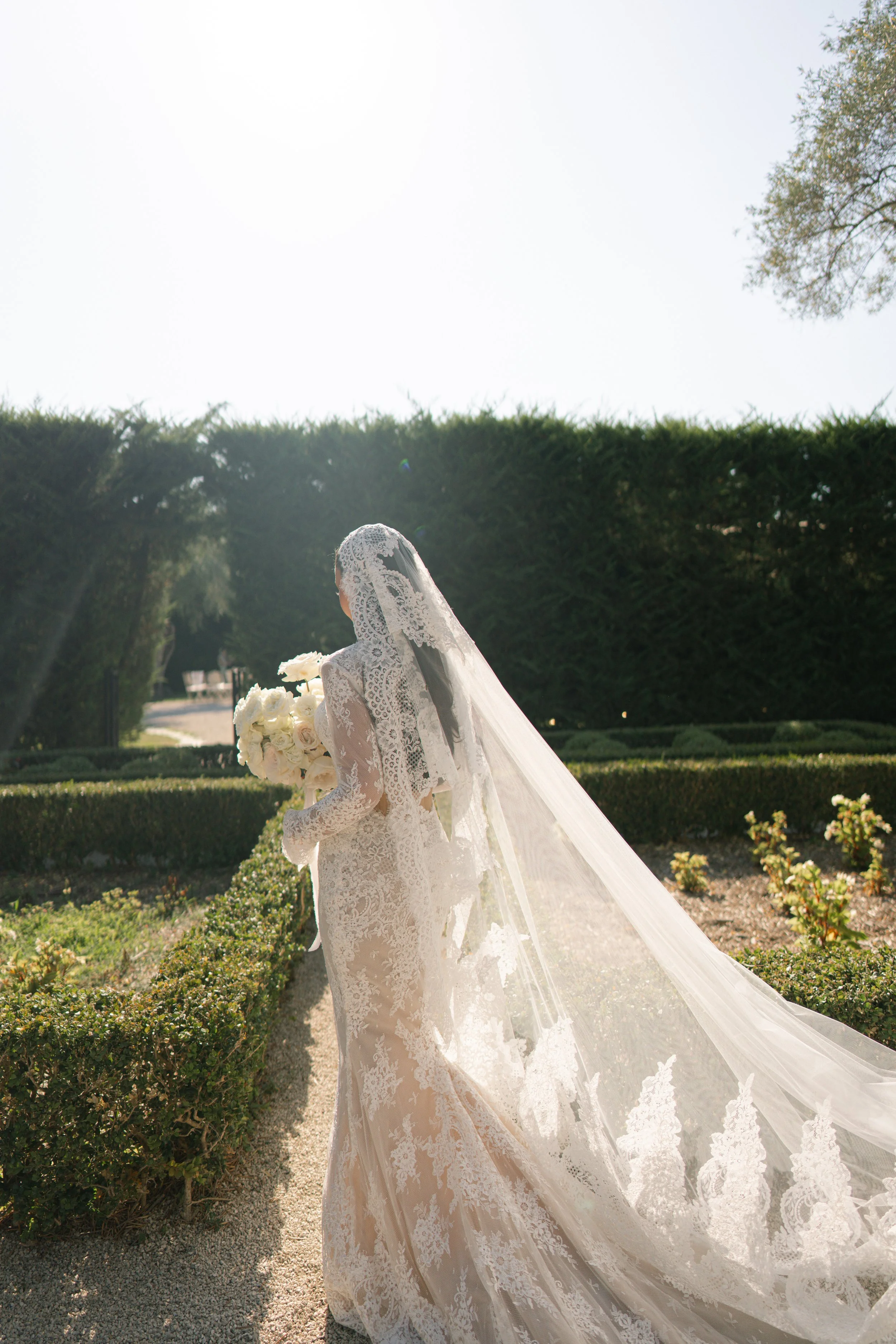 A bride wearing a lace wedding dress and veil holding a bouquet of white roses in a garden with trimmed bushes and trees, sunlight shining, and a clear sky.