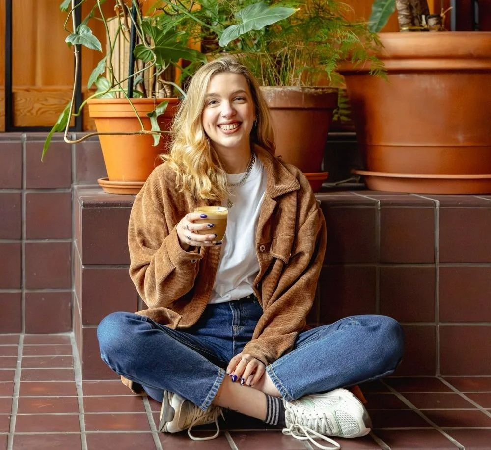 Lisa is a white woman with shoulder-length blonde hair. She is sitting on a tiled bench wearing a white t-shirt, a brown corduroy shirt, blue jeans and white trainers. She's smiling while holding a cup of coffee in one hand.
