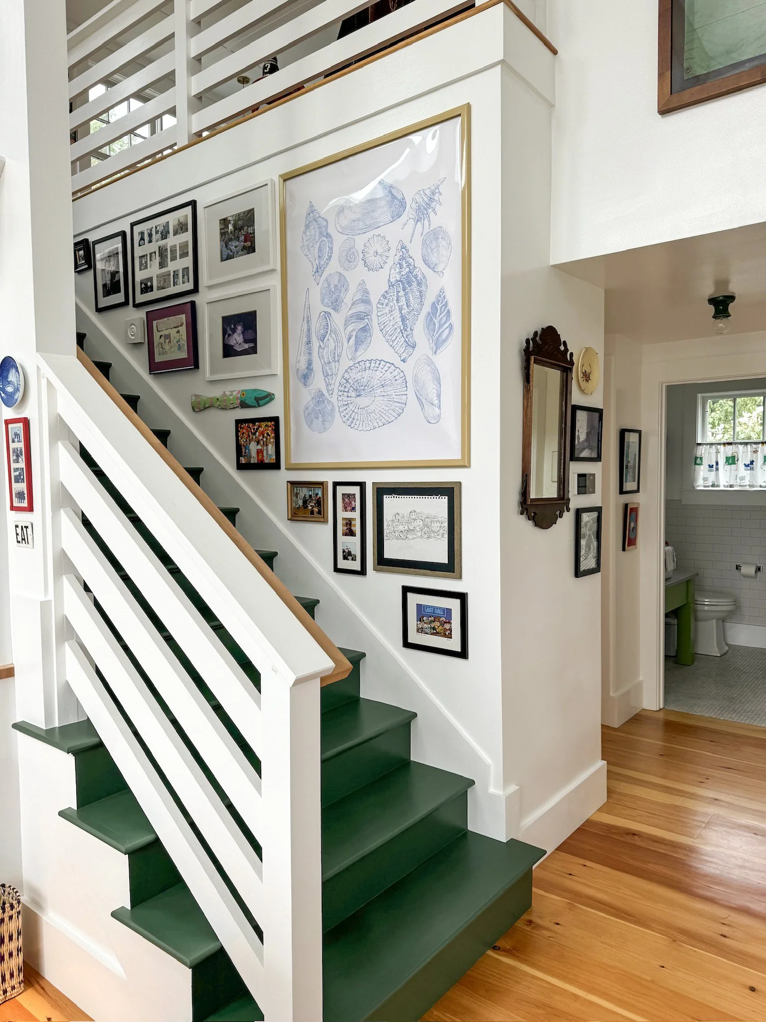Interior of a house showing a staircase with a green and white color scheme, and a gallery wall with framed photos, artwork, and decorative items. There's a bathroom visible in the background with a white toilet, a small green sink, and a window with