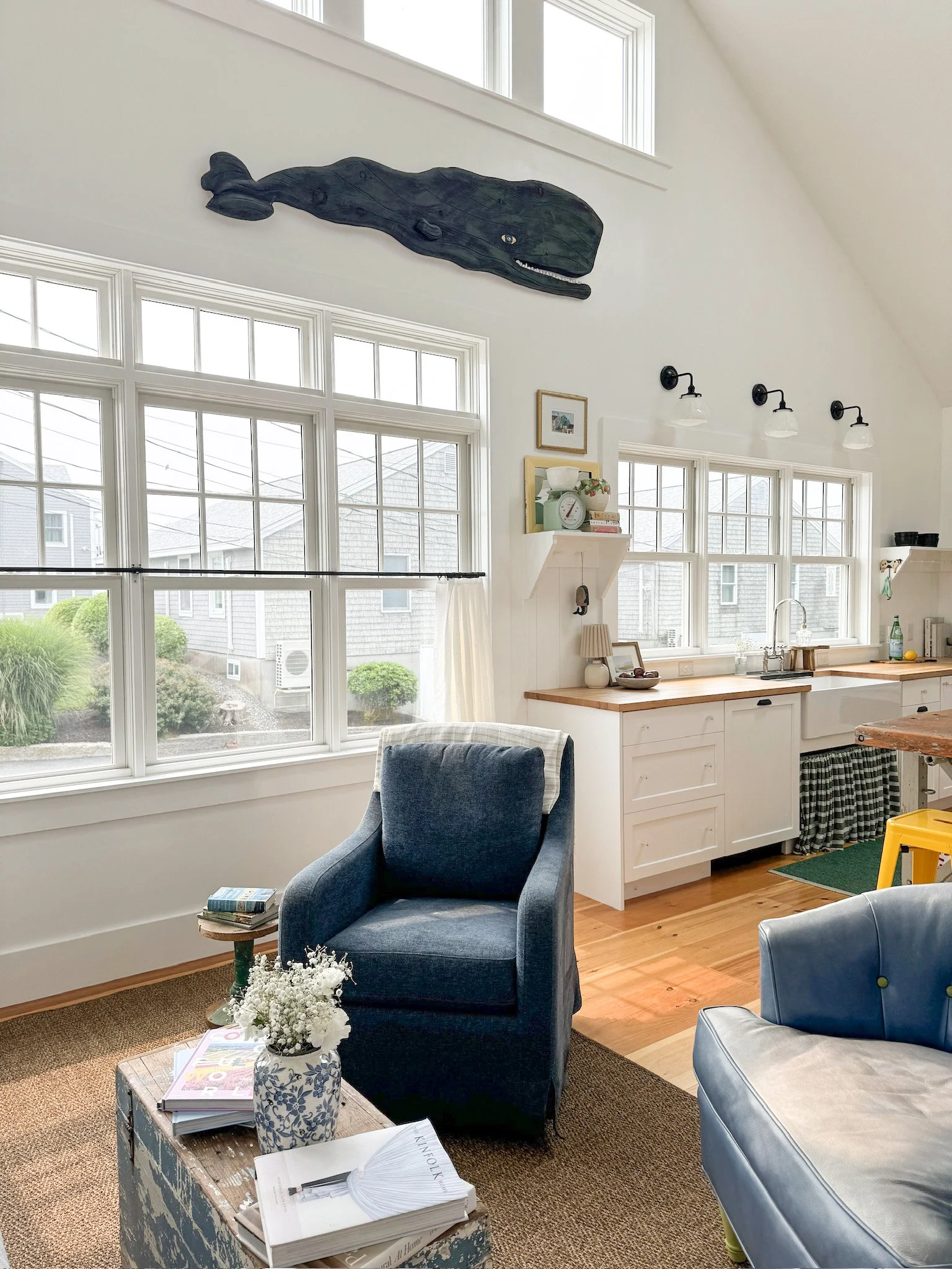 Living room with large windows, a blue armchair, a wooden coffee table with books and a vase of flowers, and white kitchen cabinets with wooden countertops.