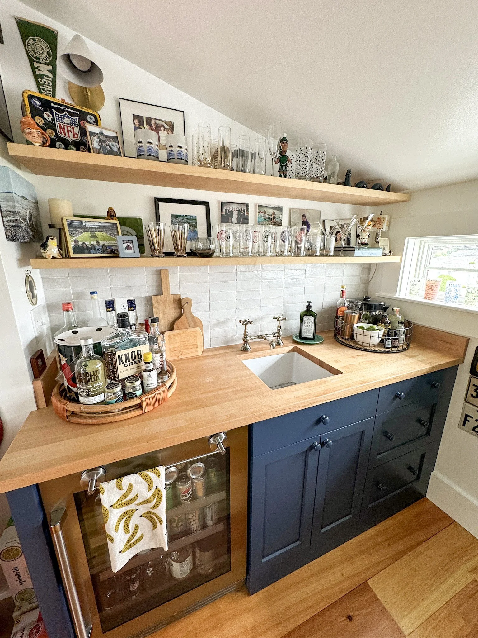 A cozy kitchen corner with a wooden countertop, navy blue cabinets, and two wooden shelves above. The shelves hold framed photos, glassware, and decorative items. On the countertop, various bottles of alcohol, a cutting board, a soap dispenser, and a