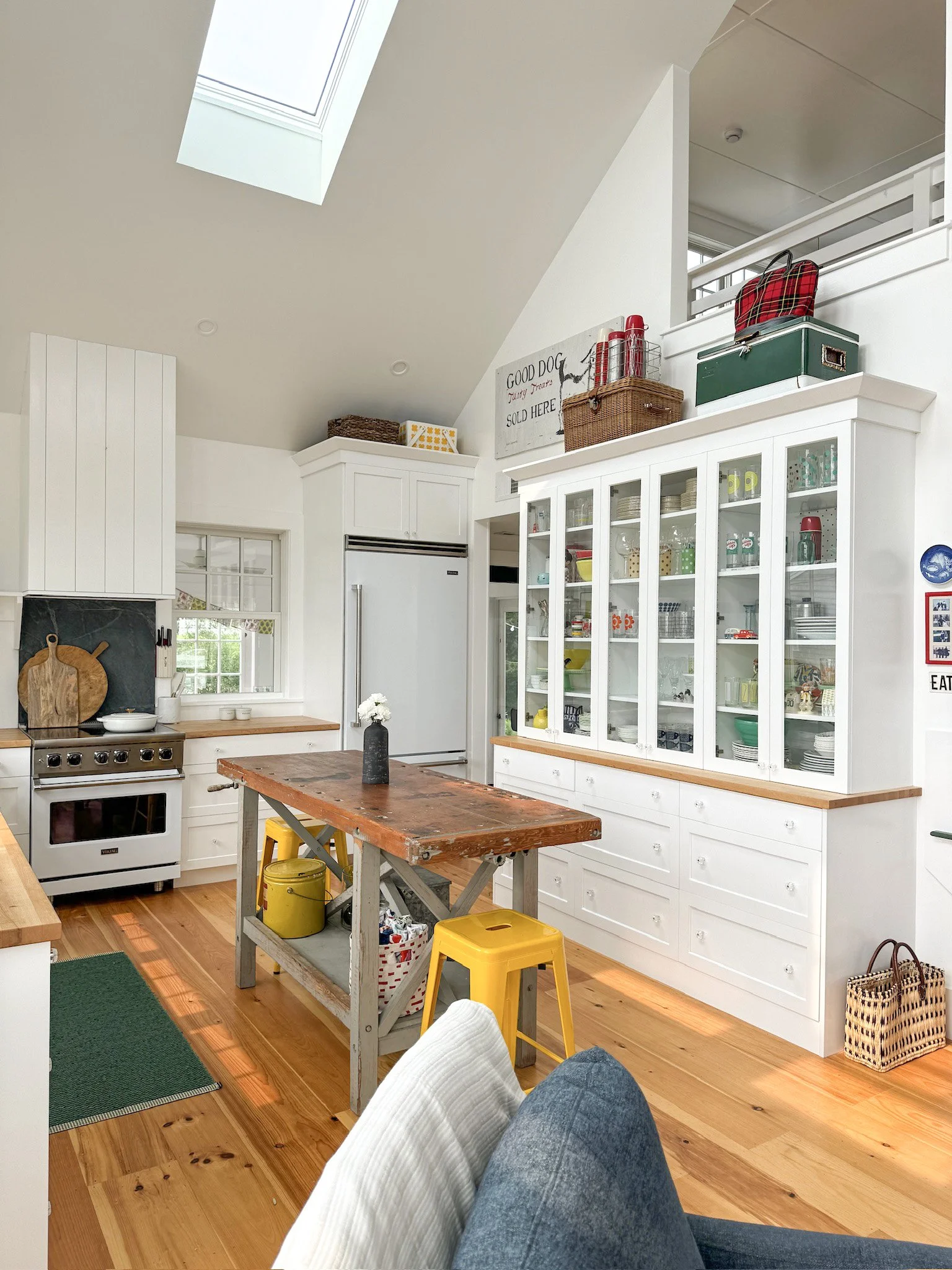 Bright kitchen with white cabinets, a wooden kitchen island with yellow stool, glass-fronted cabinet, and hardwood floors.