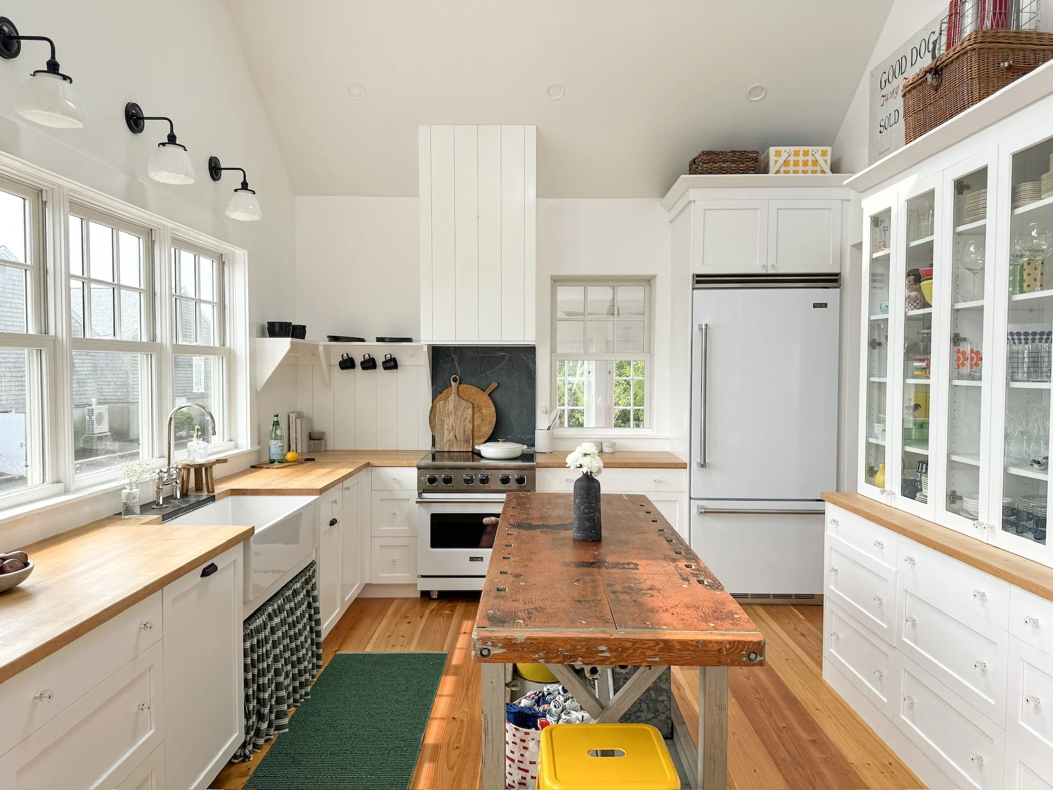 Bright kitchen with white cabinets, wooden countertops, a center island table with a worn wooden top, a large refrigerator, glass-front cabinets, and multiple windows.