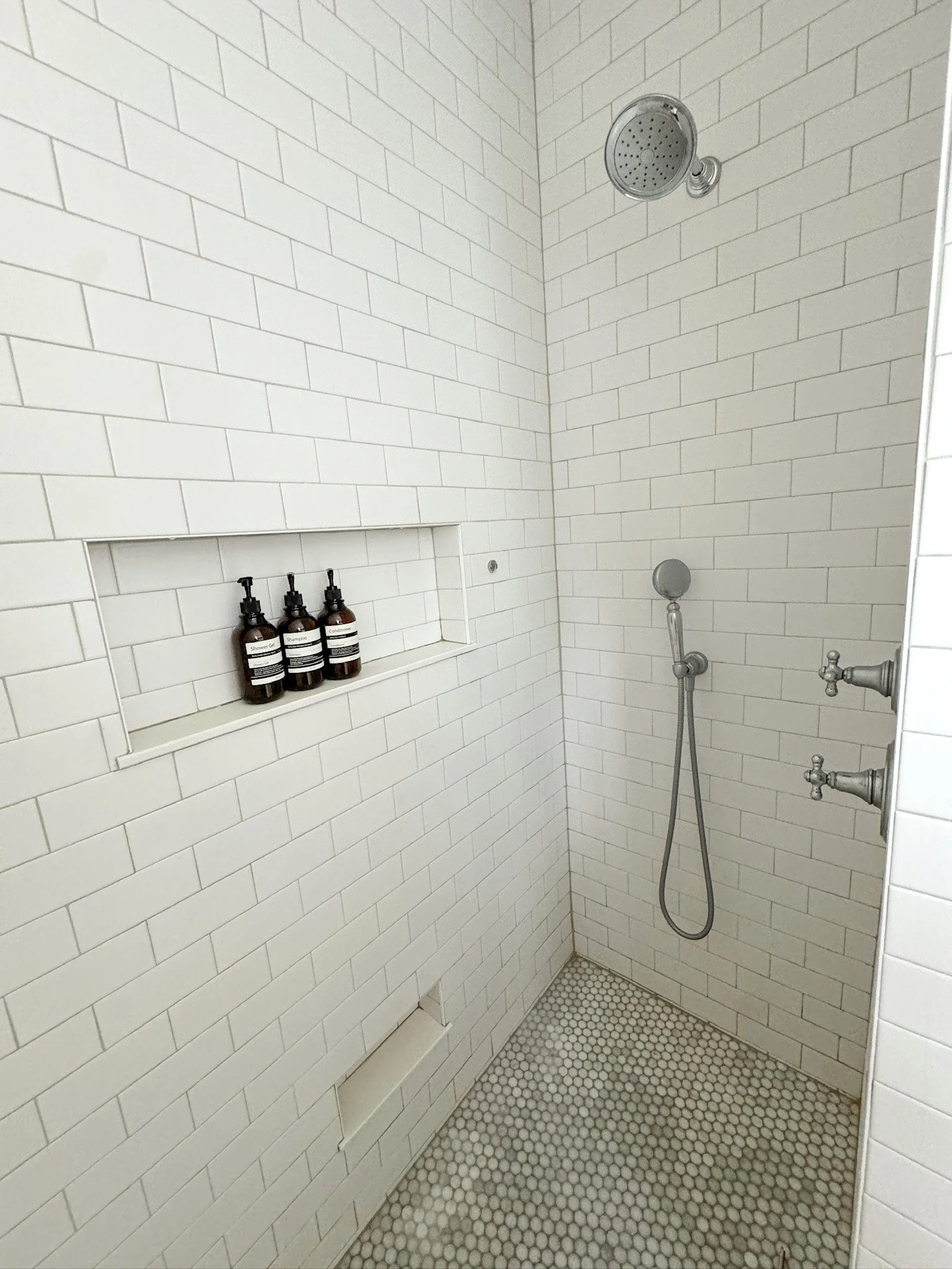 A shower with white subway tile walls, a built-in shelf with three soap dispensers, a round shower head, a handheld shower wand, and two control knobs.