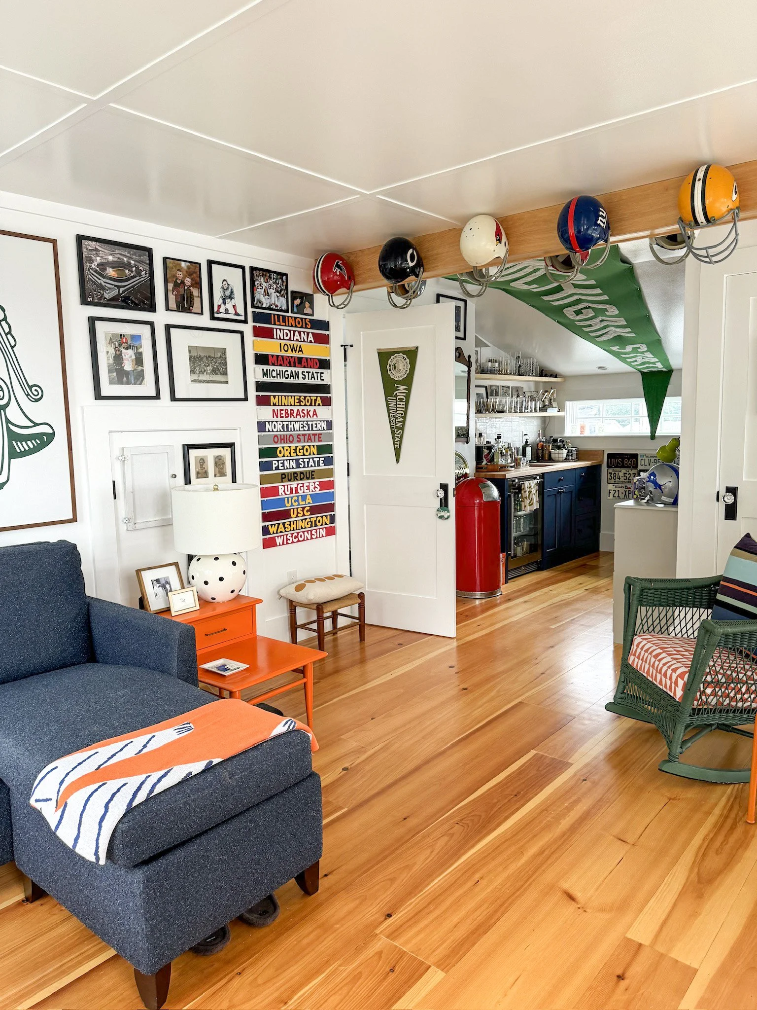 Living room with framed sports team photos and keychains, a wall with marathon and college banners, a blue armchair with a Michigan Wolverines blanket, an orange side table with a lamp, an orange stool, a green woven chair with a striped pillow, and