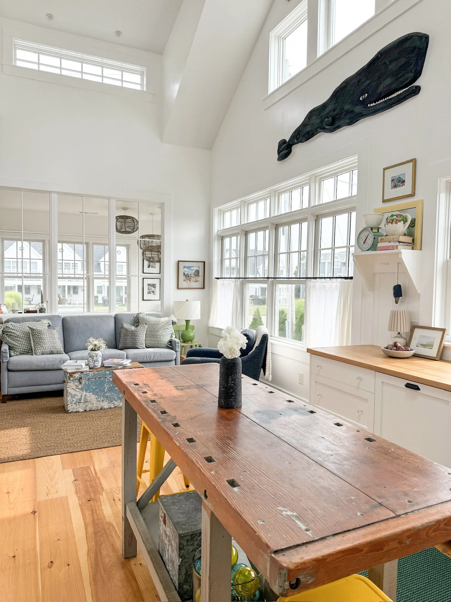 Bright living room with white walls, large windows, and a vaulted ceiling. A gray sofa with checkered pillows, a rustic wooden table, and decorative items like framed pictures, a vase with flowers, and a whale wall art are visible.