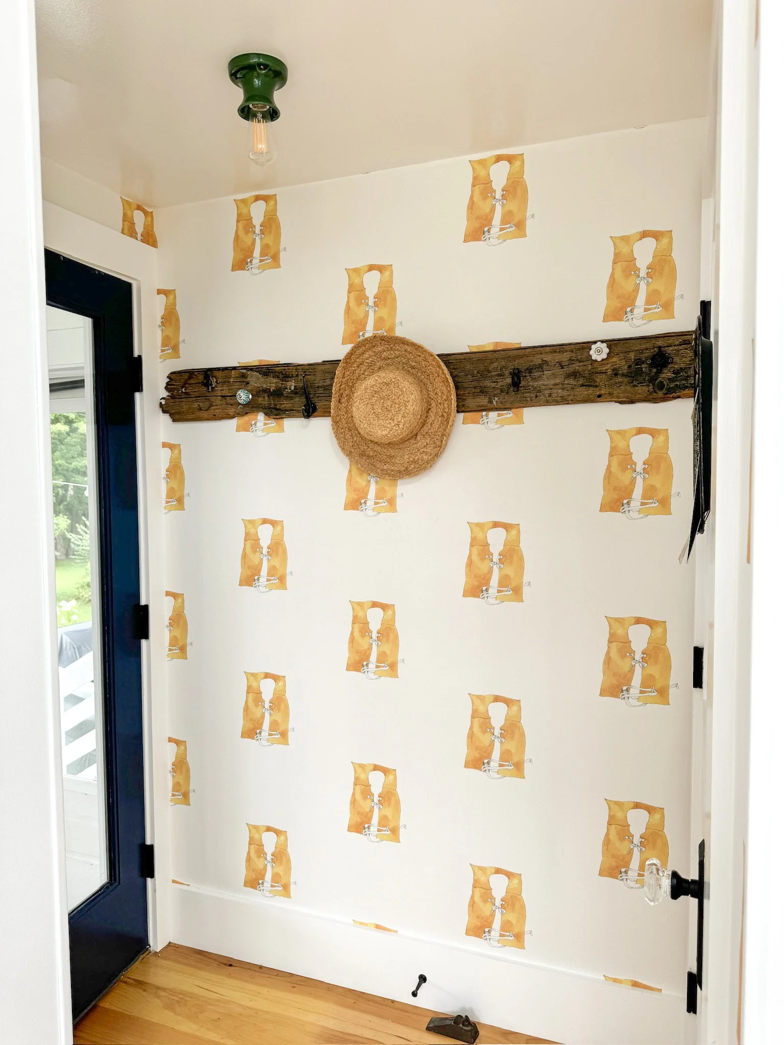 A wall with wallpaper featuring orange illustrations of light bulbs and brackets, with a wooden beam mounted across the wall. A straw hat hangs from one of the hooks on the beam, and there are small hooks and a key hanging from it.