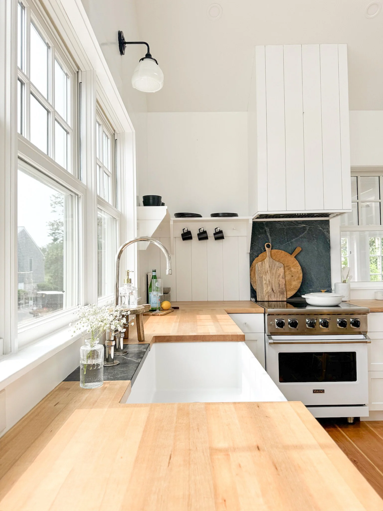 Bright kitchen with large windows, white cabinets, wooden countertops, a stainless steel stove, black utensils, and decorative cutting boards on a dark backsplash.