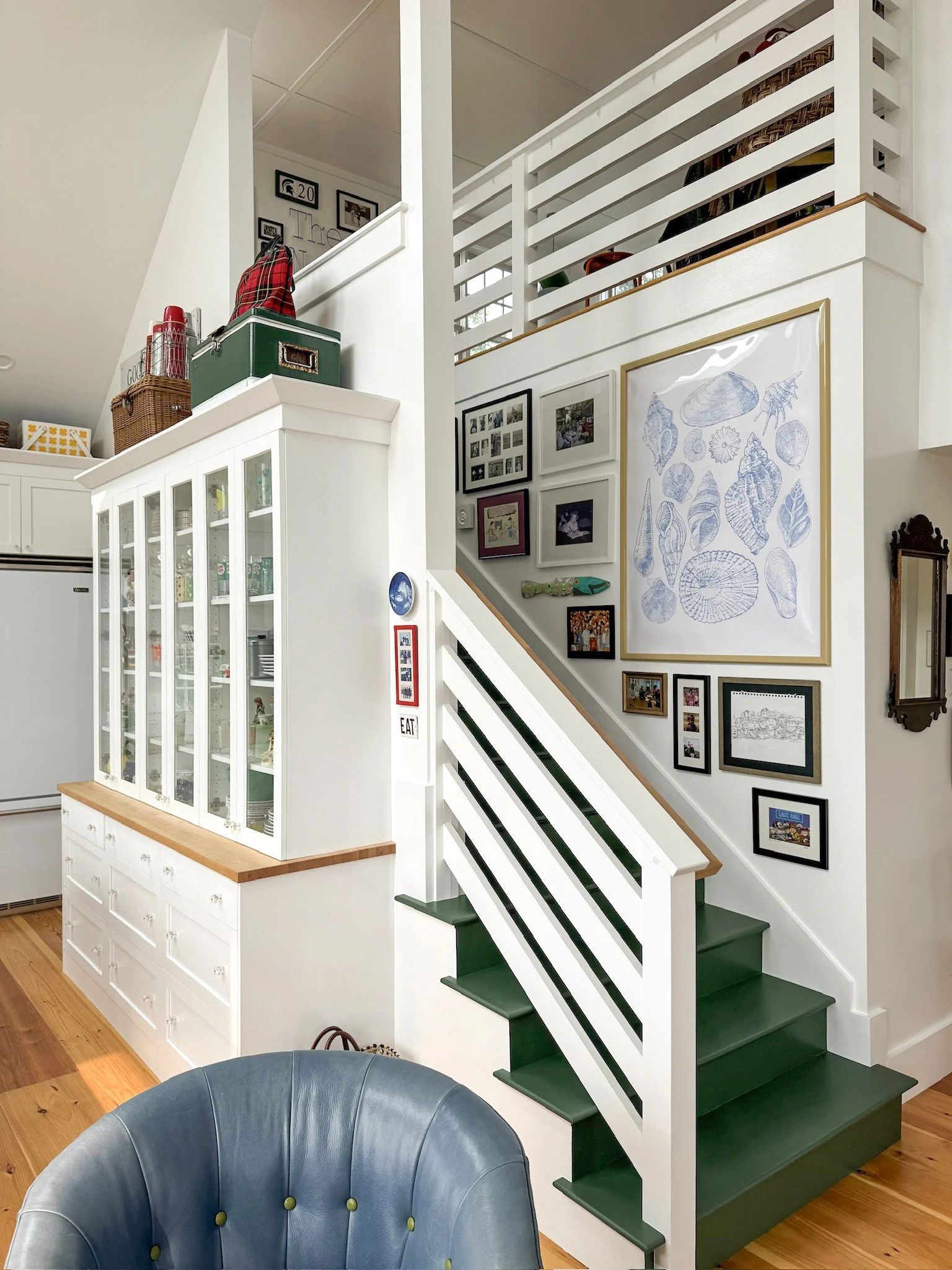 Interior view of a staircase in a home with a gallery wall of framed pictures and blue seashell drawing, a colorful fish sculpture, and a green bench at the base of the stairs.
