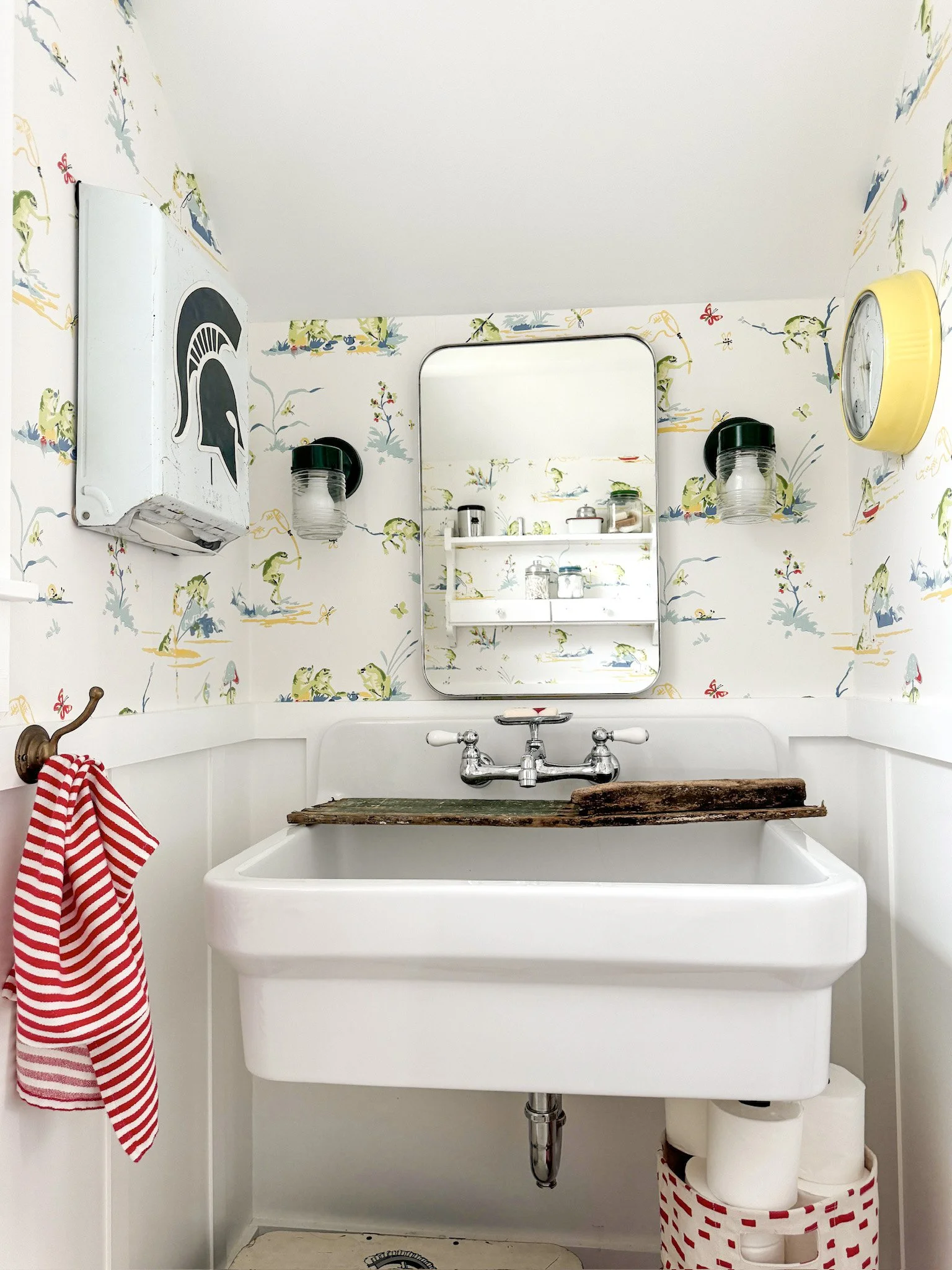 A vintage farmhouse bathroom with a white sink, a wooden plank on top, a small mirror, and patterned wallpaper featuring frogs and plants. There is a red and white striped towel on a hook, and a basket holding toilet paper rolls beneath the sink. Two