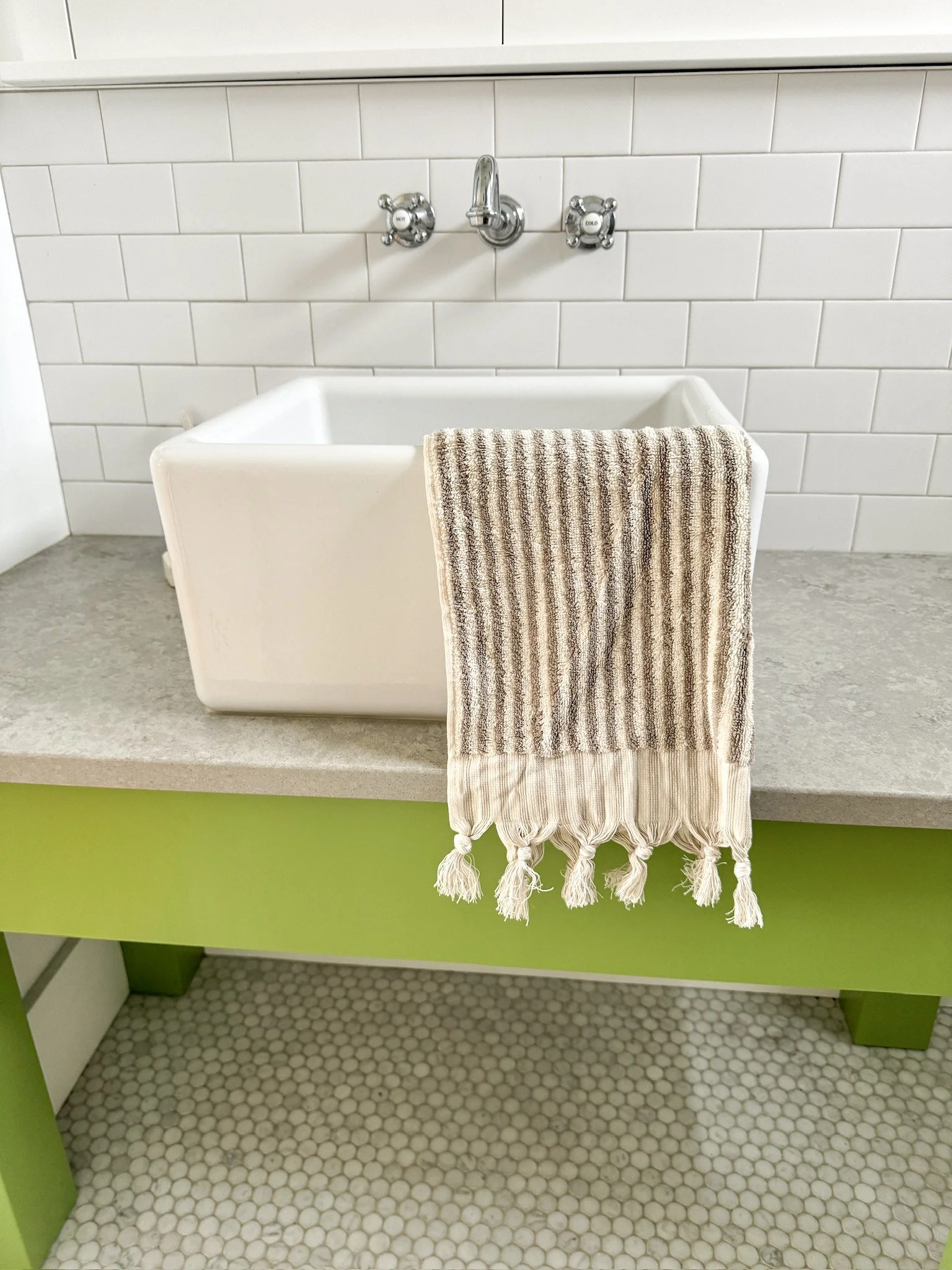 A white laundry sink with a beige and brown striped towel hanging over it, in a room with white subway tiles on the wall and green and grey cabinetry.
