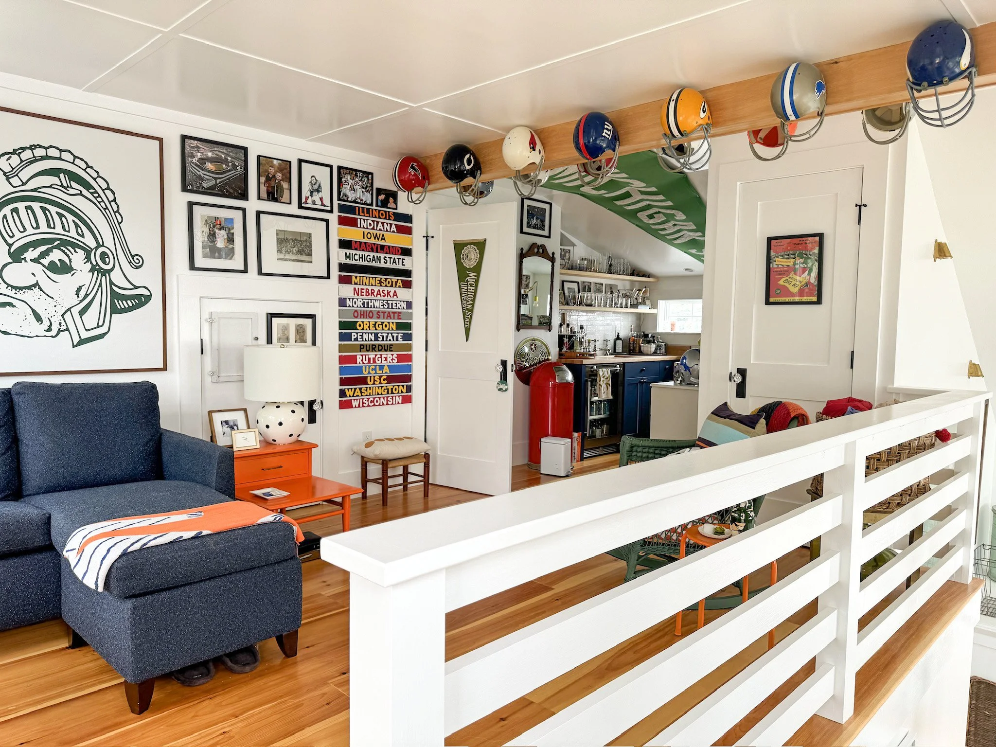 Living room decorated with football helmets hanging from a wooden beam, sports posters, framed photos, and a Michigan State banner, with a dark blue armchair, an orange side table, a lamp, and a white railing dividing the space.
