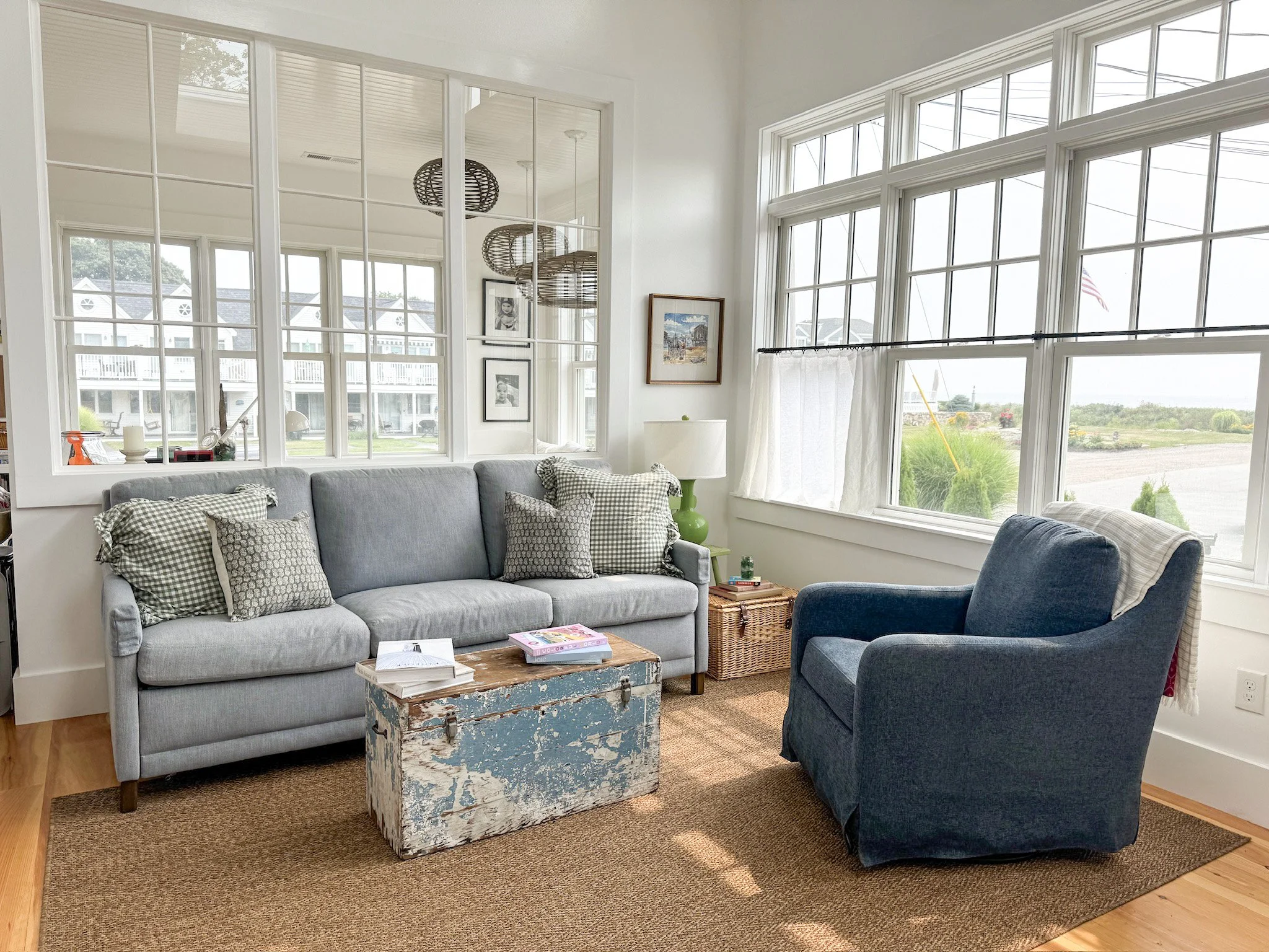 Living room with a grey sofa, a blue armchair, wooden coffee table, wicker side table, green lamp, large windows, and outdoor view.
