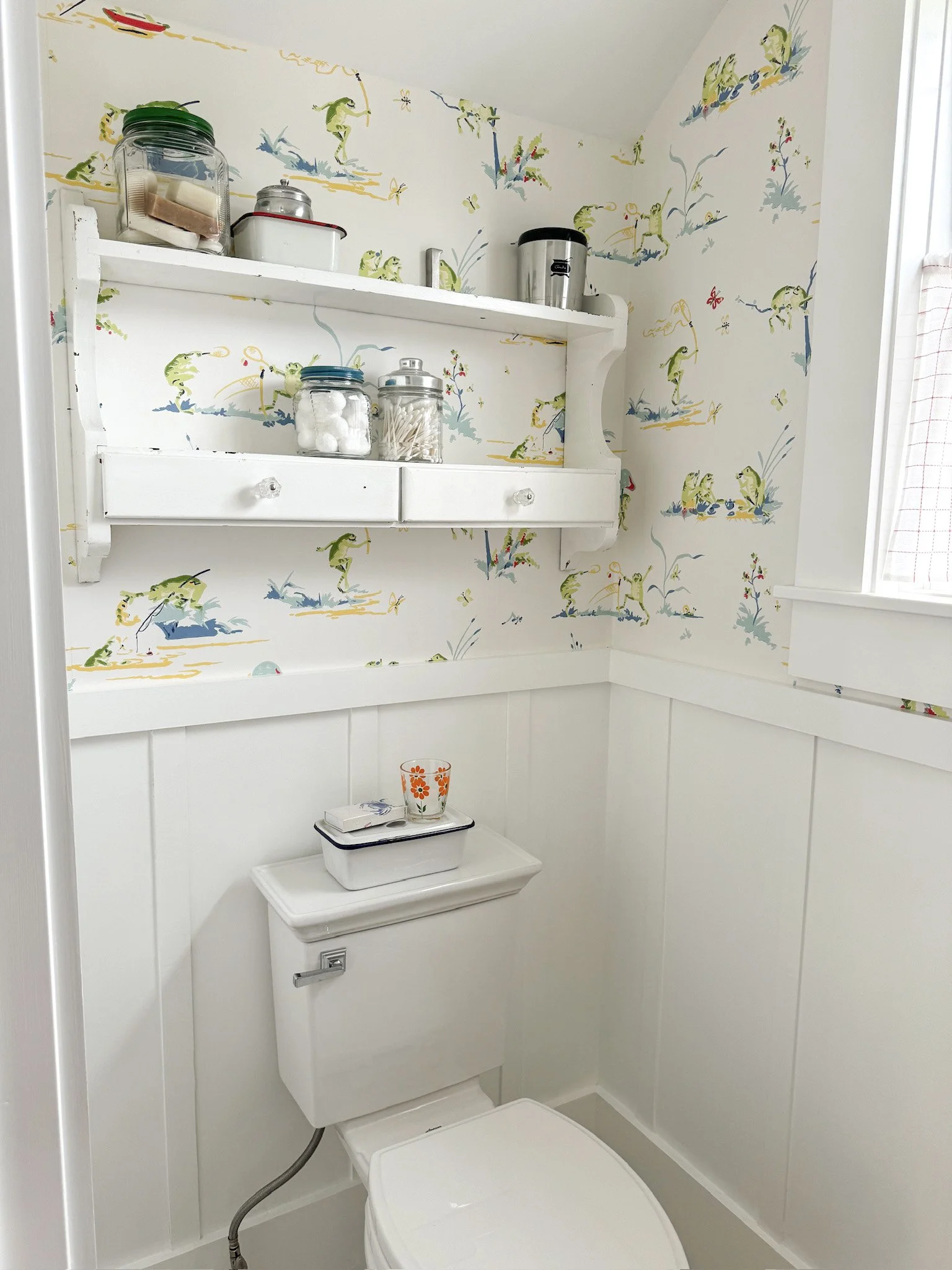 A small bathroom with wallpaper featuring frogs, flowers, and nature scenes. A white toilet with a small shelf on top holding a container with orange flower design, a box of tissues, and a small book. A white wall-mounted shelf with two drawers above