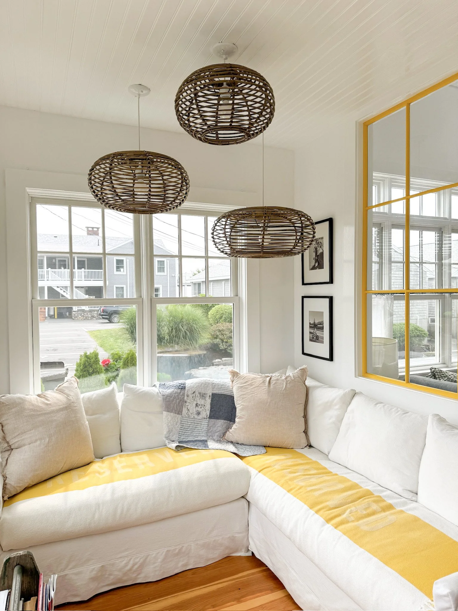 Bright living room with large windows, a white sectional sofa adorned with beige and gray pillows, hanging wicker pendant lights, and framed black-and-white photos on the wall.