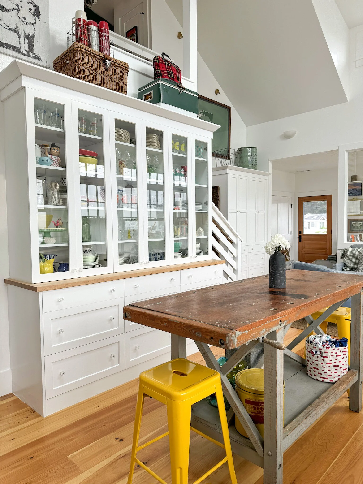 A spacious kitchen with a rustic wooden table, a yellow stool, and a white cabinet with glass doors displaying dishes and kitchenware. Items are stored on top of the cabinet and on a nearby shelf. The kitchen has wood flooring and a door with a windo