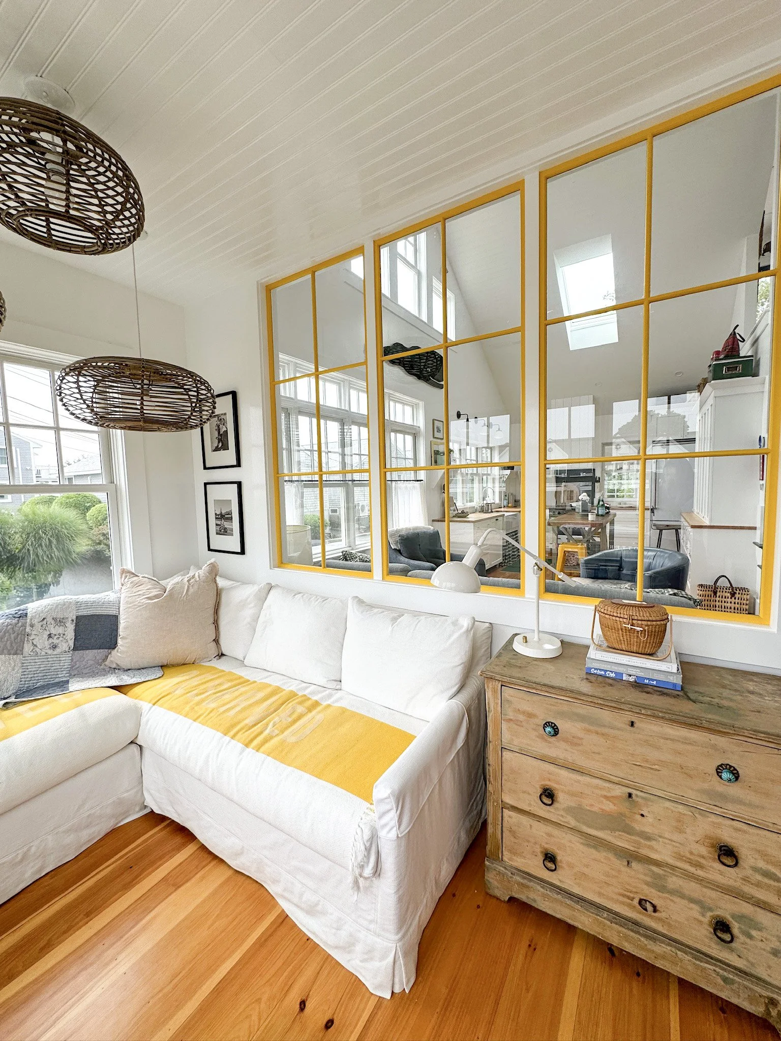 Living room with white sofa, yellow throw blanket, and mixed pillows; wooden dresser with books and basket; yellow-framed window mirror reflecting kitchen and dining area; natural light from multiple windows and skylights.