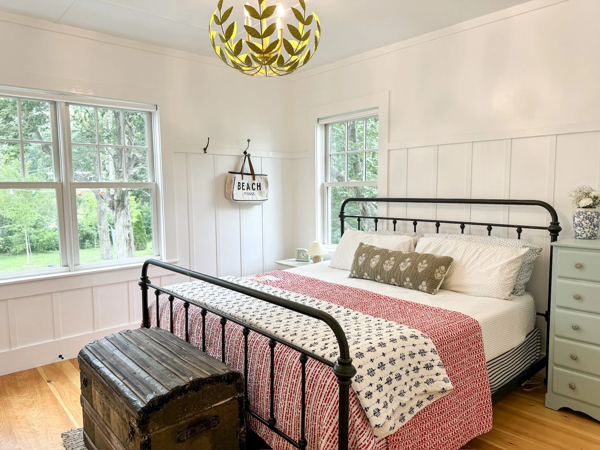 A cozy bedroom with white walls and wooden floors, featuring a black metal bed with white and patterned bedding, a vintage chest at the foot of the bed, a mint green dresser with a floral vase, a small nightstand with a lamp, and large windows showin
