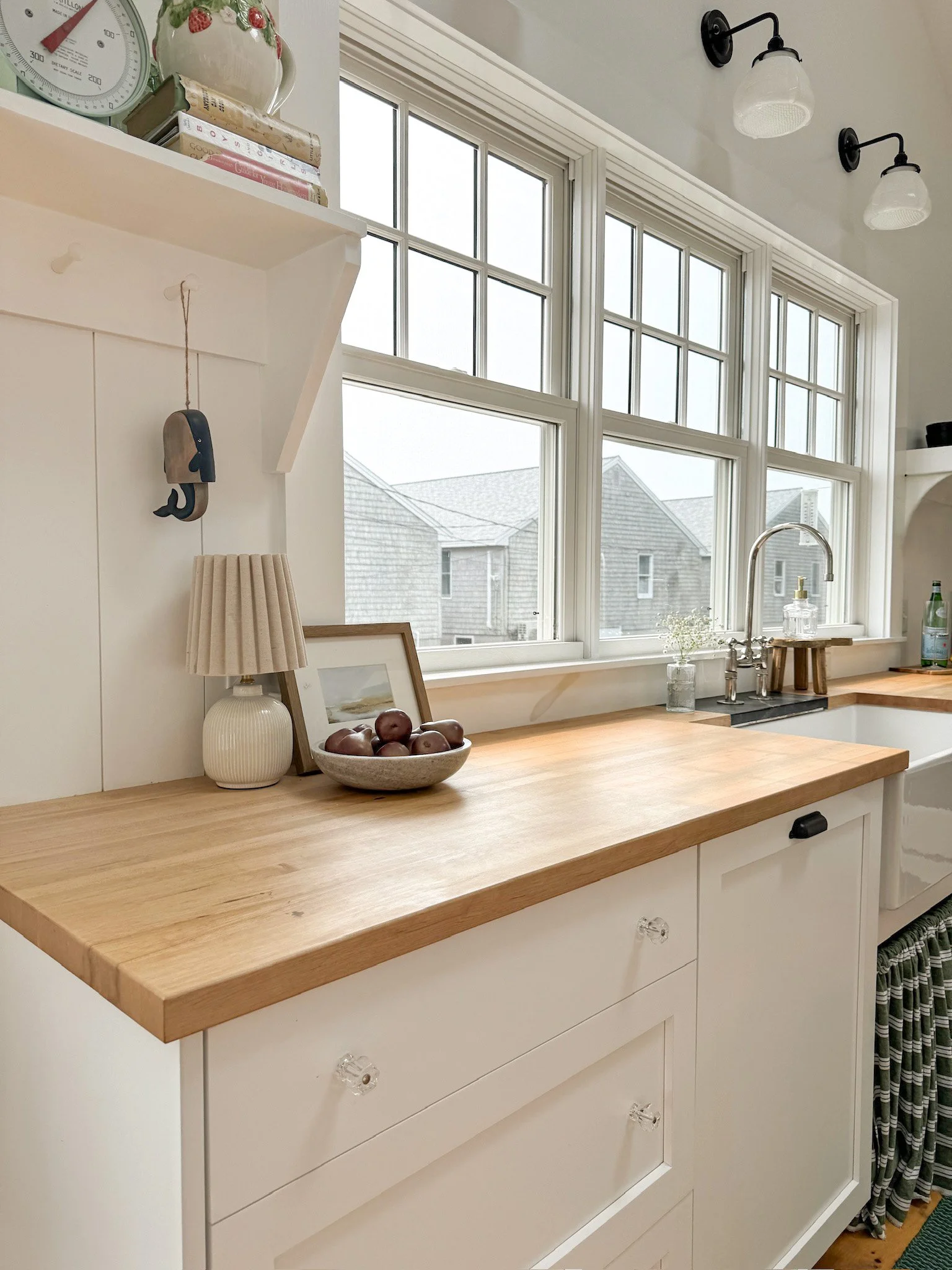 Kitchen with white cabinets, light wood countertop, and large windows. Decor includes a small lamp, a bowl of apples, framed photo, and glass bottles near the sink.