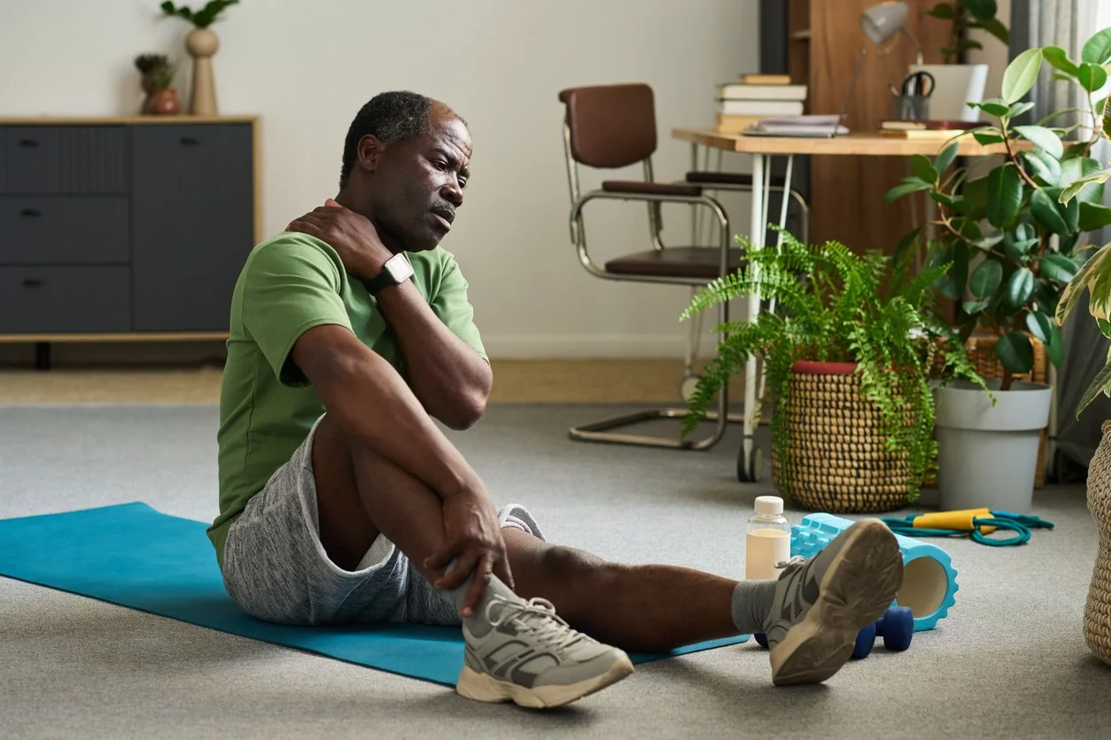 Middle-aged man sitting on a mat at home holding his neck in discomfort, representing stiffness and uncertainty when starting exercise after 40