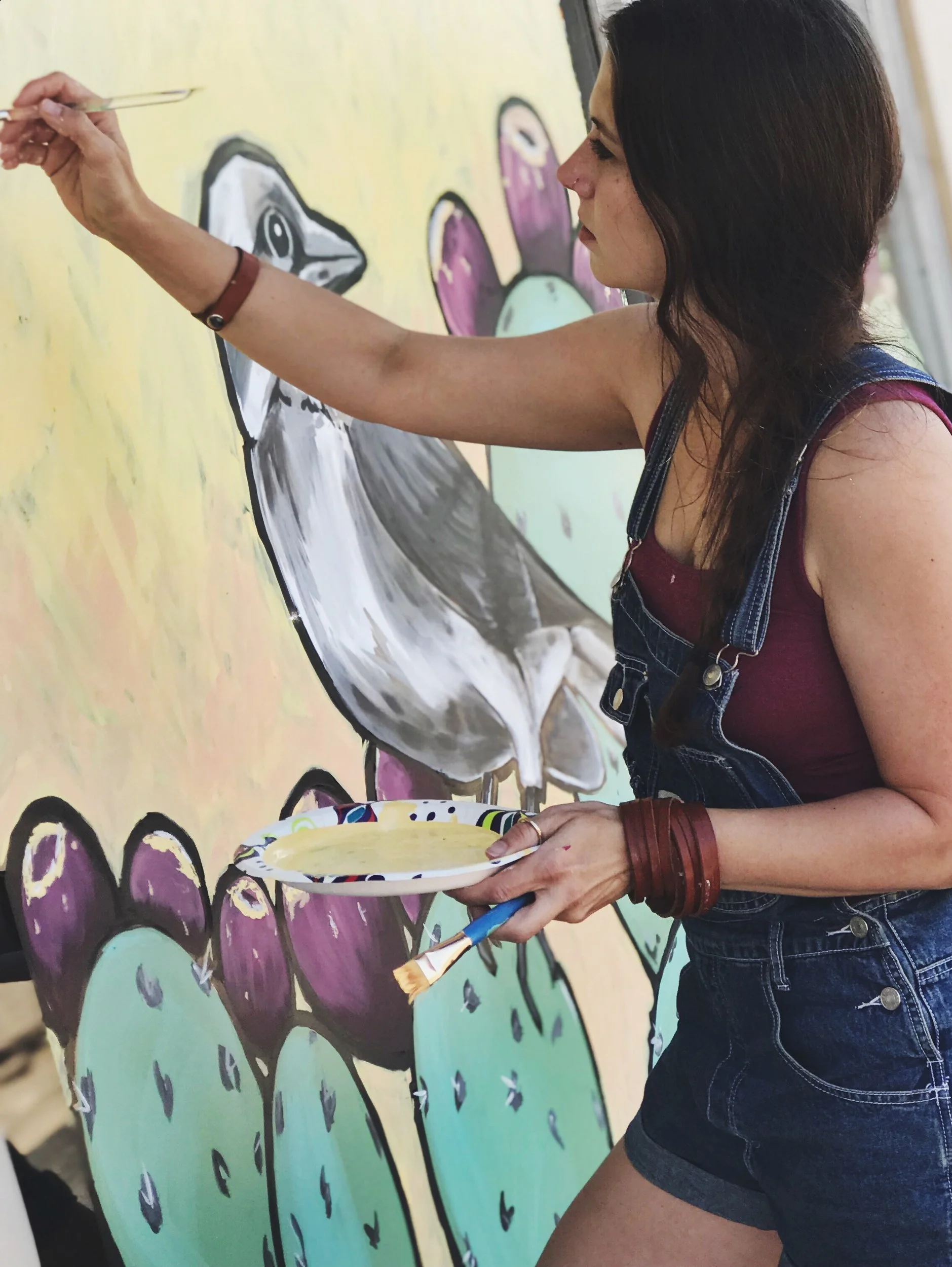 A woman painting a colorful mural that features stylized flowers with purple petals and green stems on a yellow background.