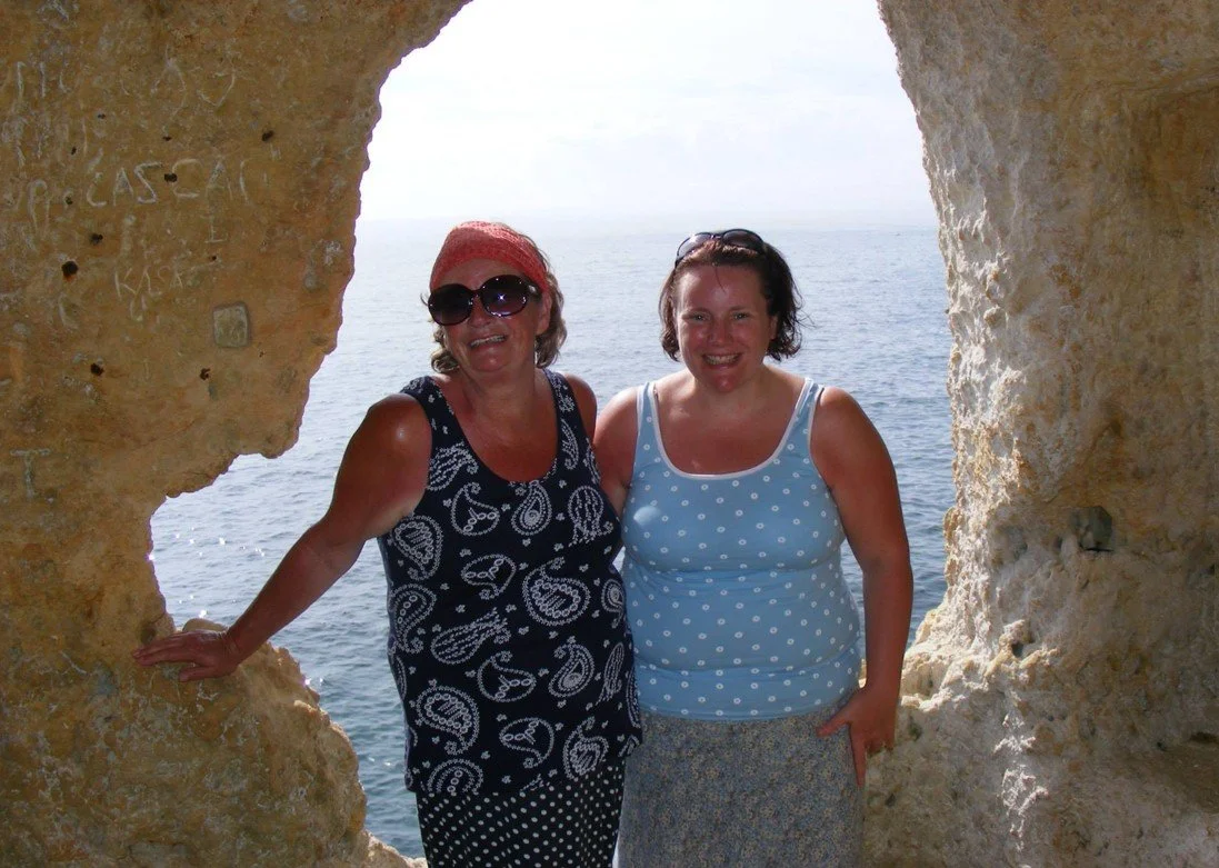 Tracy and her mum standing in the gap of a cave in Carvoeiro, Portugal, the sea behind them. Tracy is wearing a patterned blue top and skirt, her mum a patterned black top and skirt.