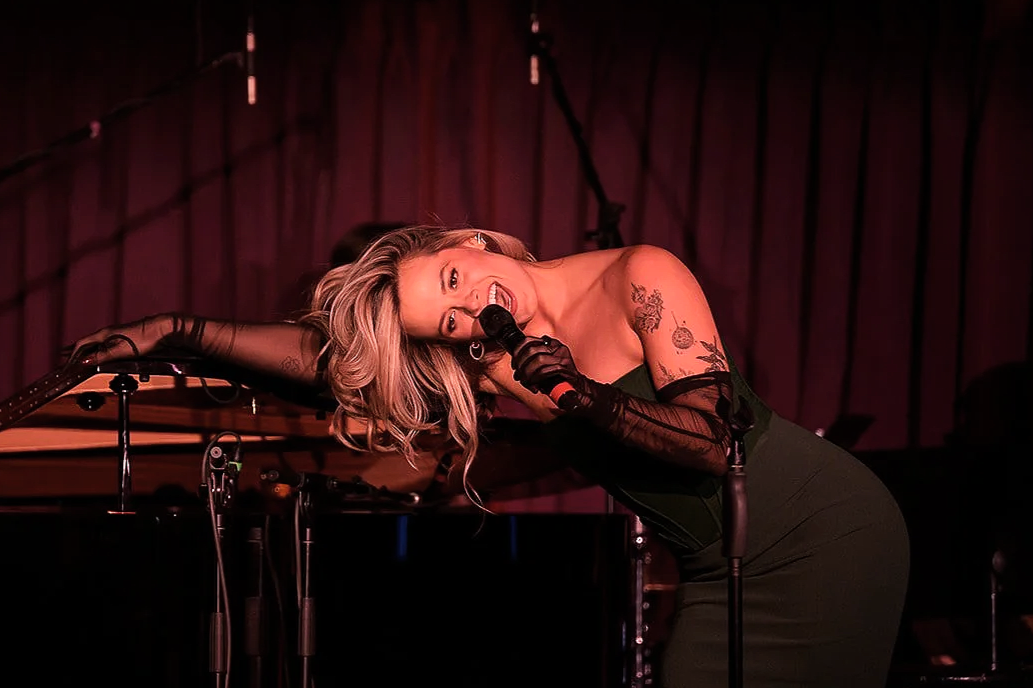 Hailie leans across a piano, head sitting on her shoulder, as she sings into a microphone