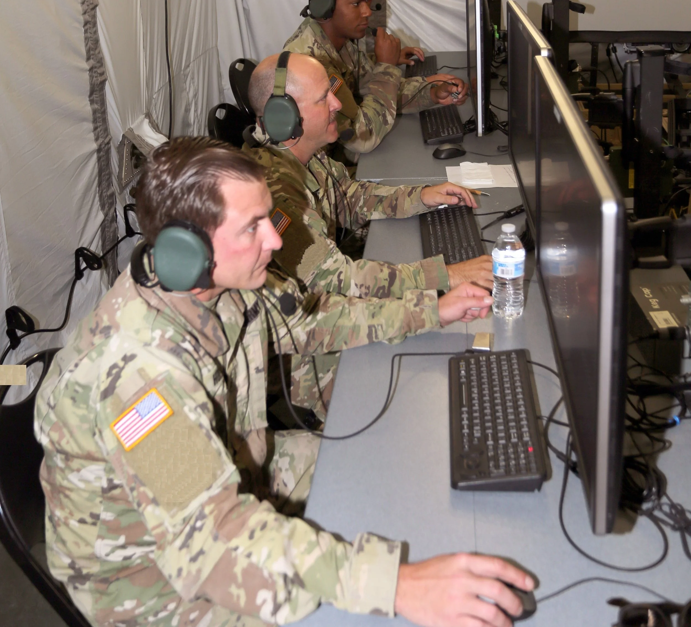 U.S. Army soldiers wearing headsets operating IBCS workstations inside a military command environment