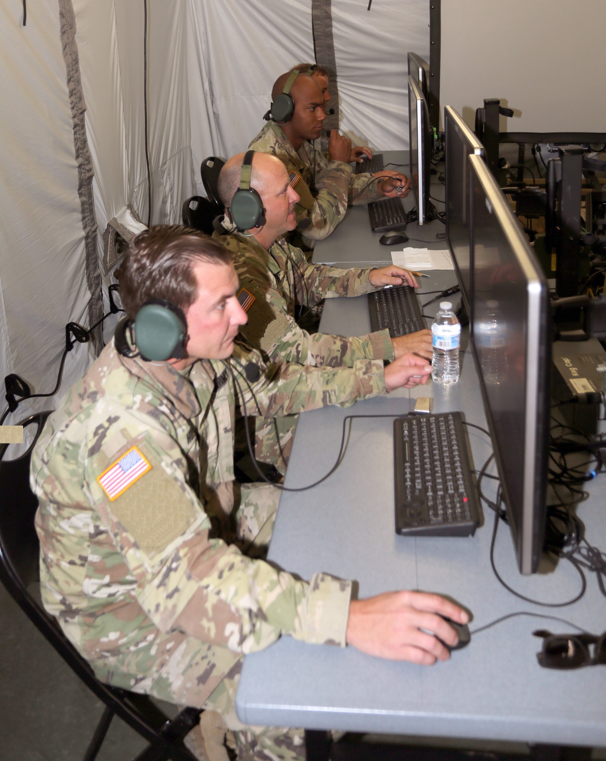 U.S. Army soldiers wearing headsets operating IBCS workstations inside a military command environment