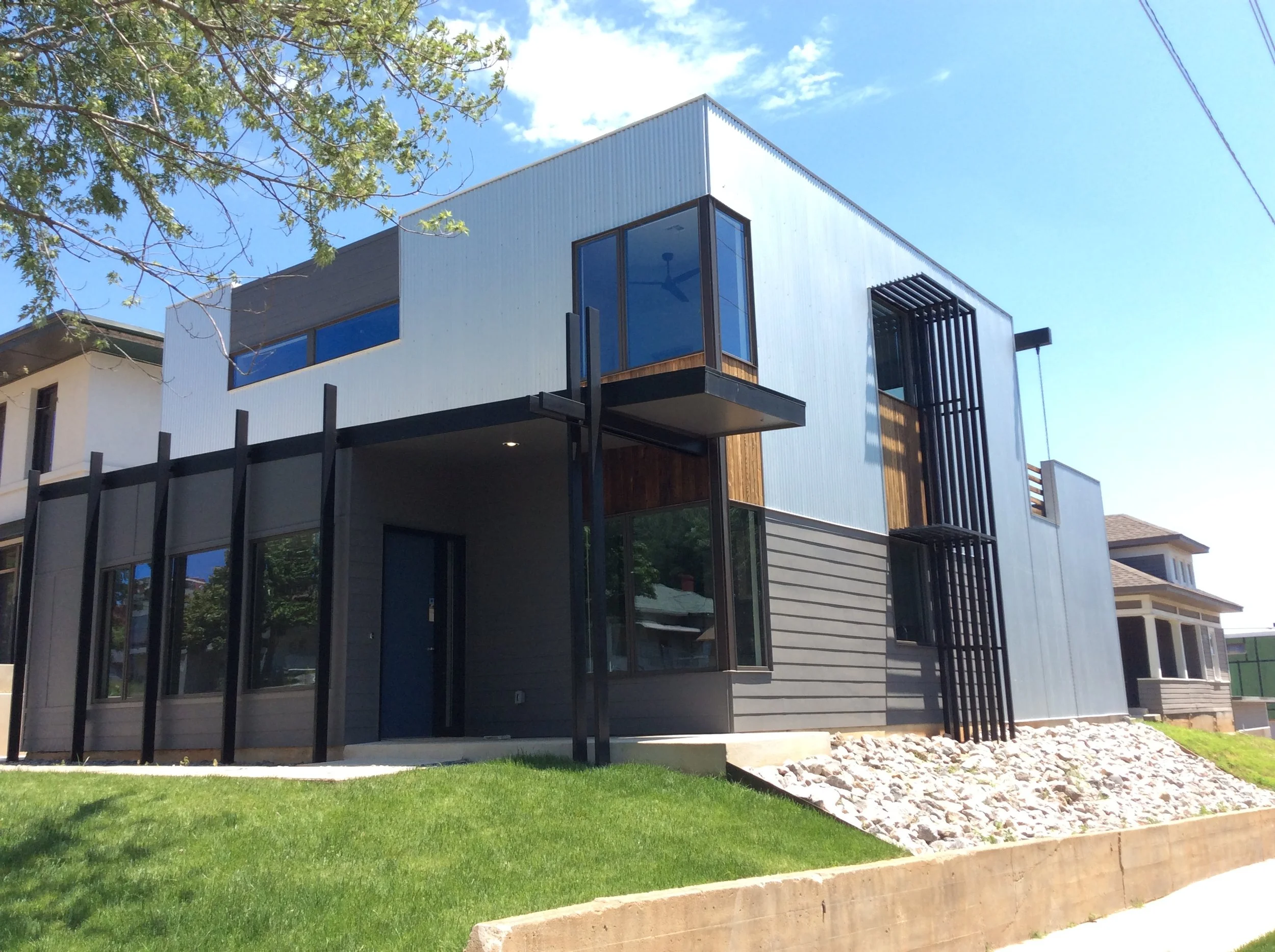 Modern two-story house with black and gray exterior, large windows, a small balcony, and a landscaped yard with grass and rocks.