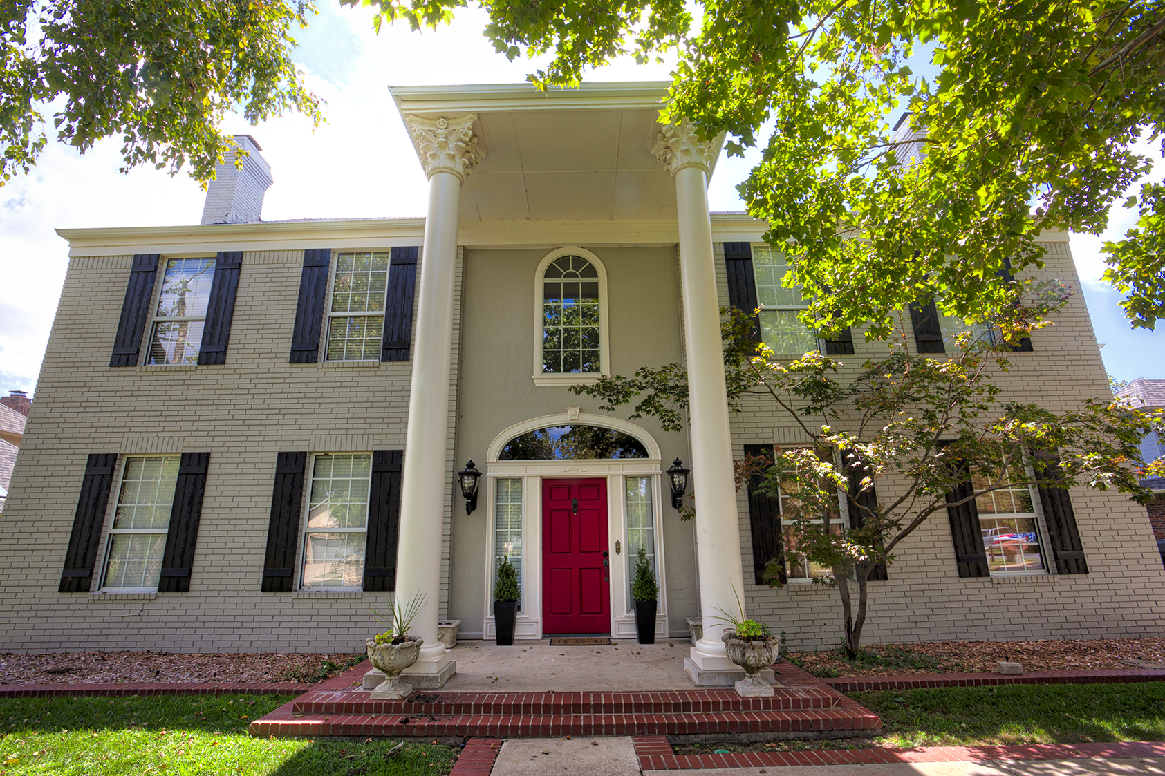 A two-story house with a red front door, white brick exterior, black shutters on the windows, two large white columns at the entrance, and lush green trees around the front yard.