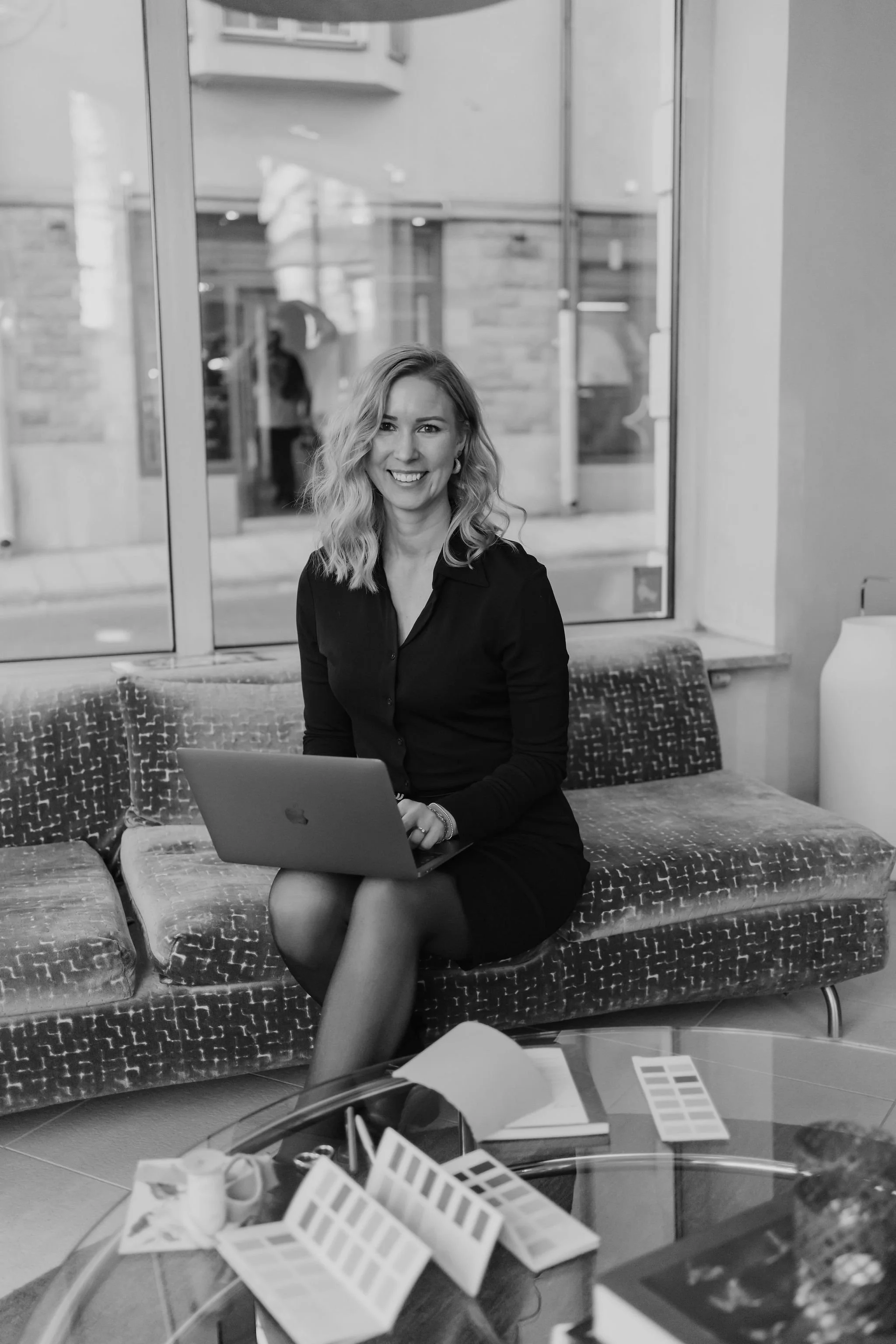 A woman with wavy hair, smiling, sitting on a patterned couch with a laptop on her lap inside a cafe or lounge.
