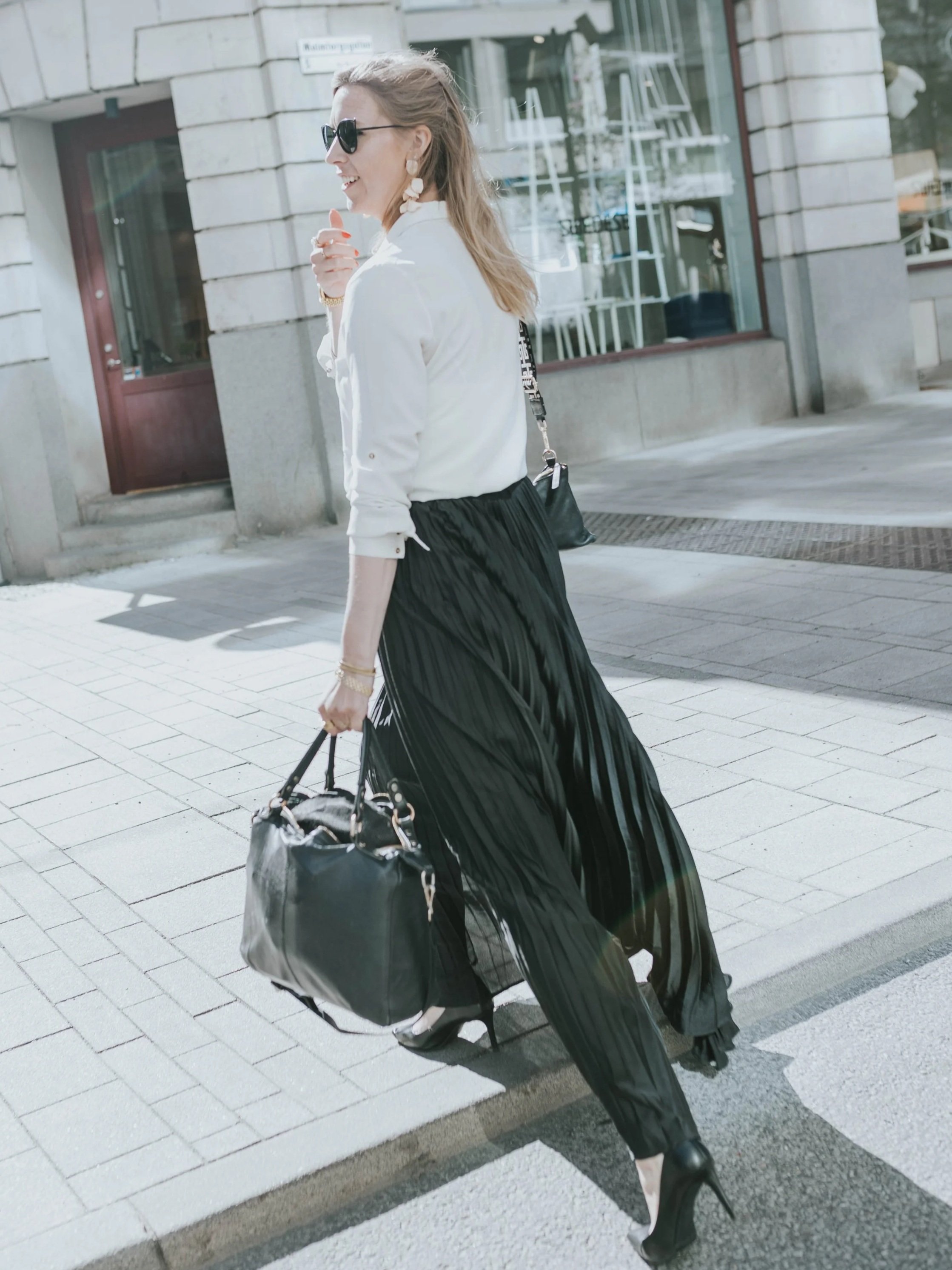 A woman walking on a city sidewalk, wearing sunglasses, a white shirt, a black pleated skirt, and high heels, carrying a black handbag.