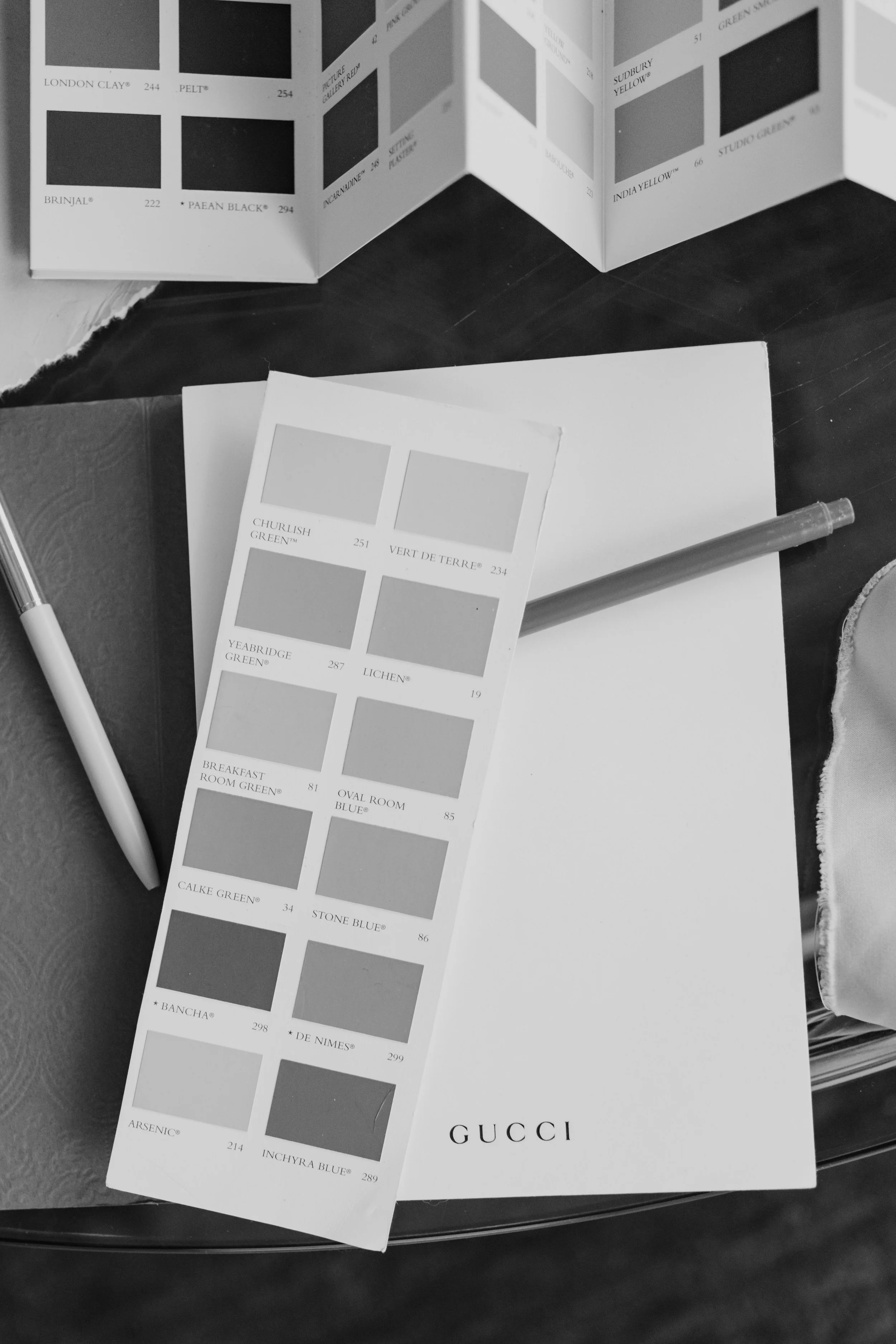 A black-and-white photo of paint color swatches and a color sample card on a dark surface, with a pen nearby. The color swatches feature shades like green, blue, and gray, with brand names and numbers.