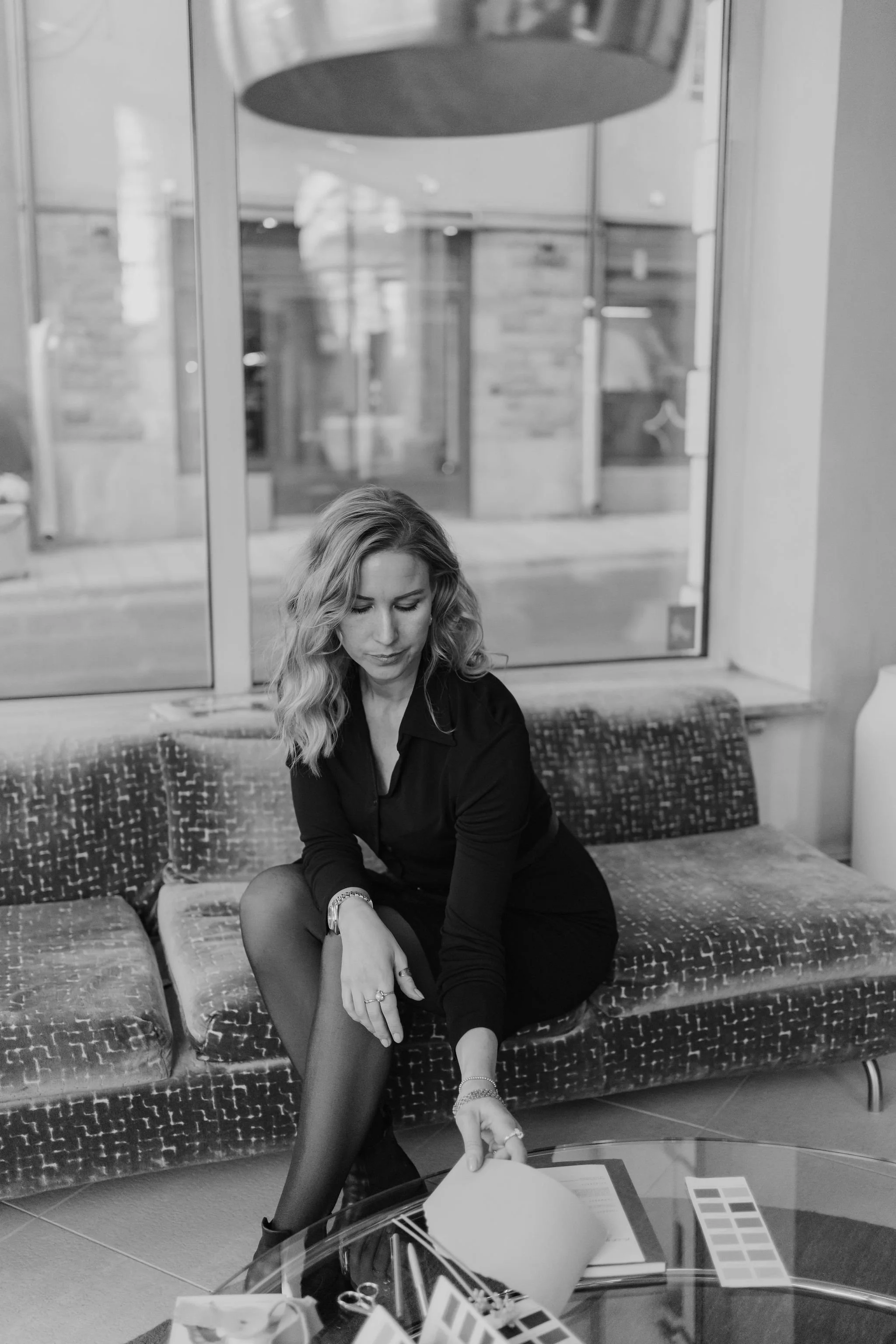 A young woman with wavy blonde hair is sitting on a patterned couch in a modern indoor space. She is looking down, adjusting her black pantyhose or tights. The background includes large glass windows and a wooden wall. A clear coffee table with vario