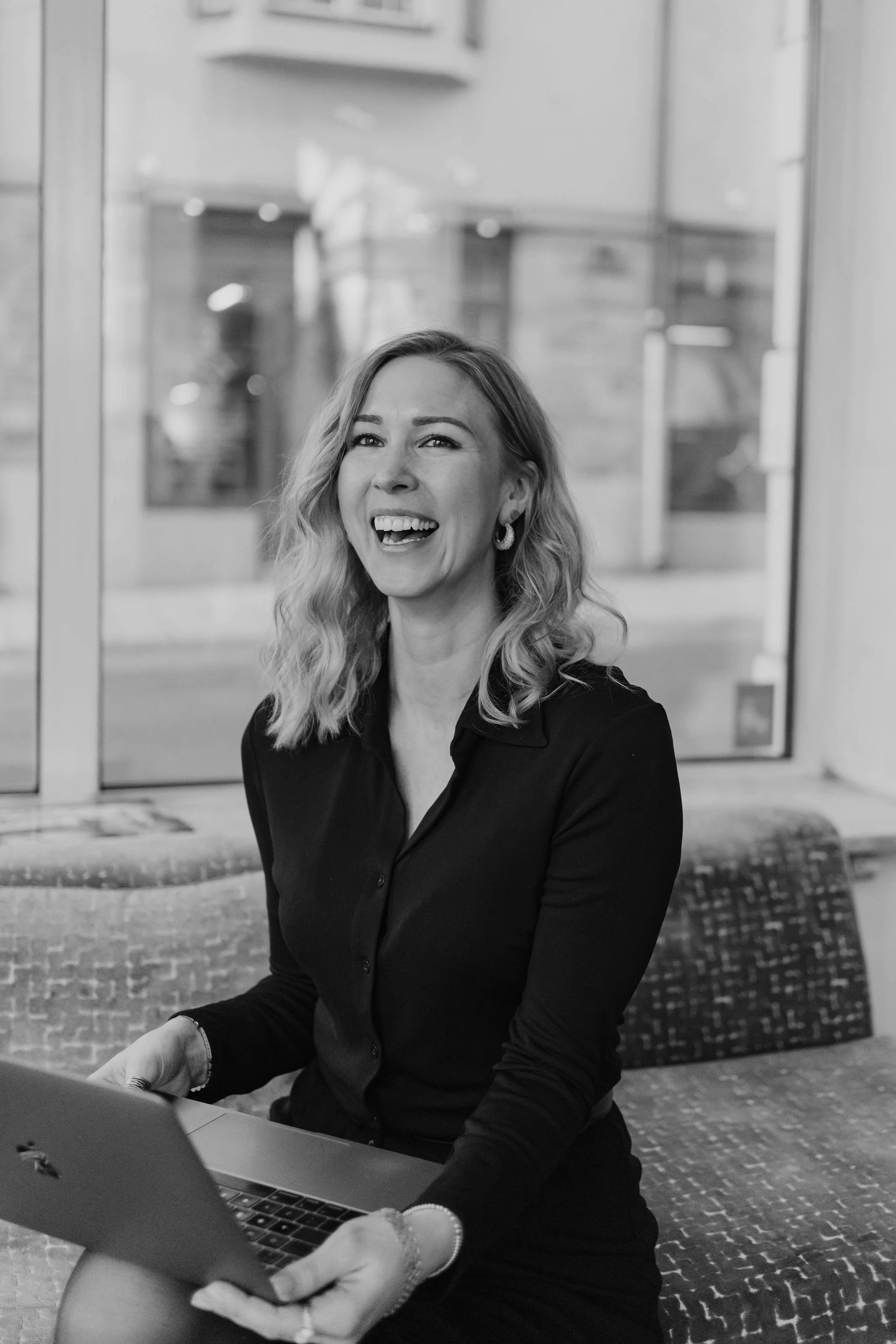 A woman with wavy hair smiling and looking at a tablet while sitting outside near a brick building with glass doors.