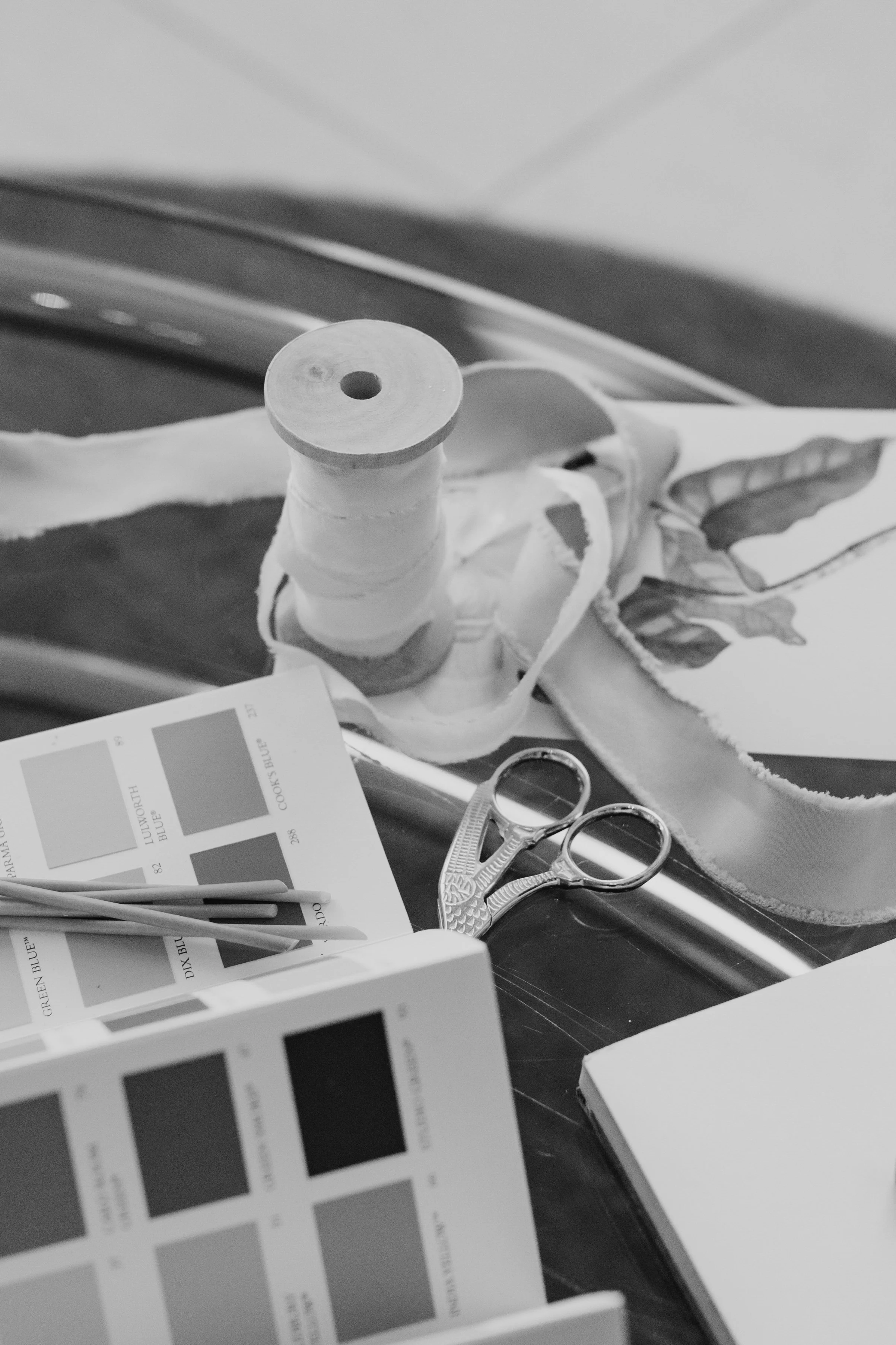 A black and white photo of a workspace with fabric, color swatch cards, scissors, and a spool of thread.