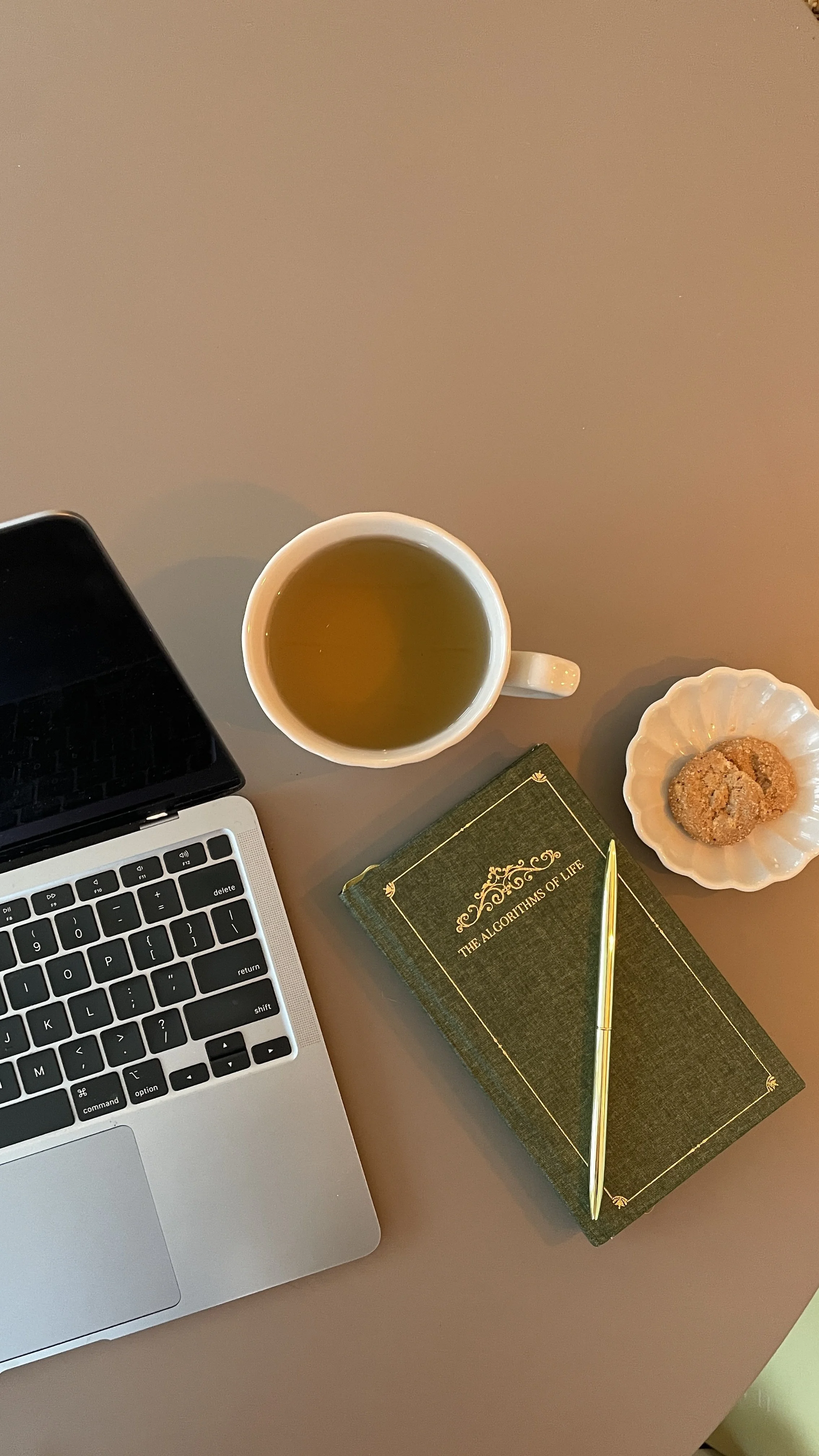 Laptop, cup of tea, book titled 'The Algorithms of Life,' a small dish with cookies, and a gold pen on a pink surface.