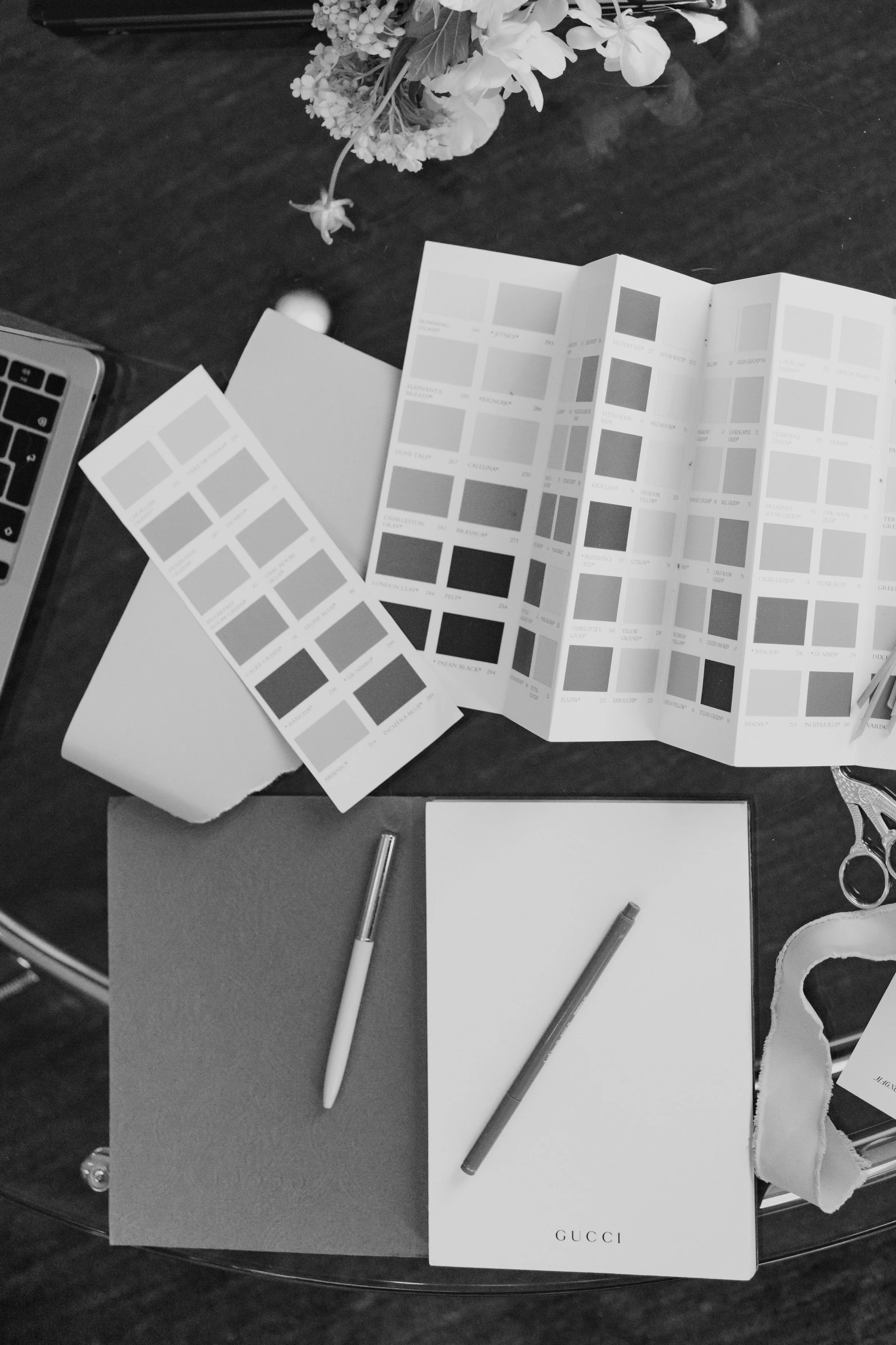 Black and white photo of a workspace with a floral arrangement, paint color sample swatches, two pens, a Gucci branded notebook, scissors, a pair of glasses, and part of a laptop on a dark wooden surface.