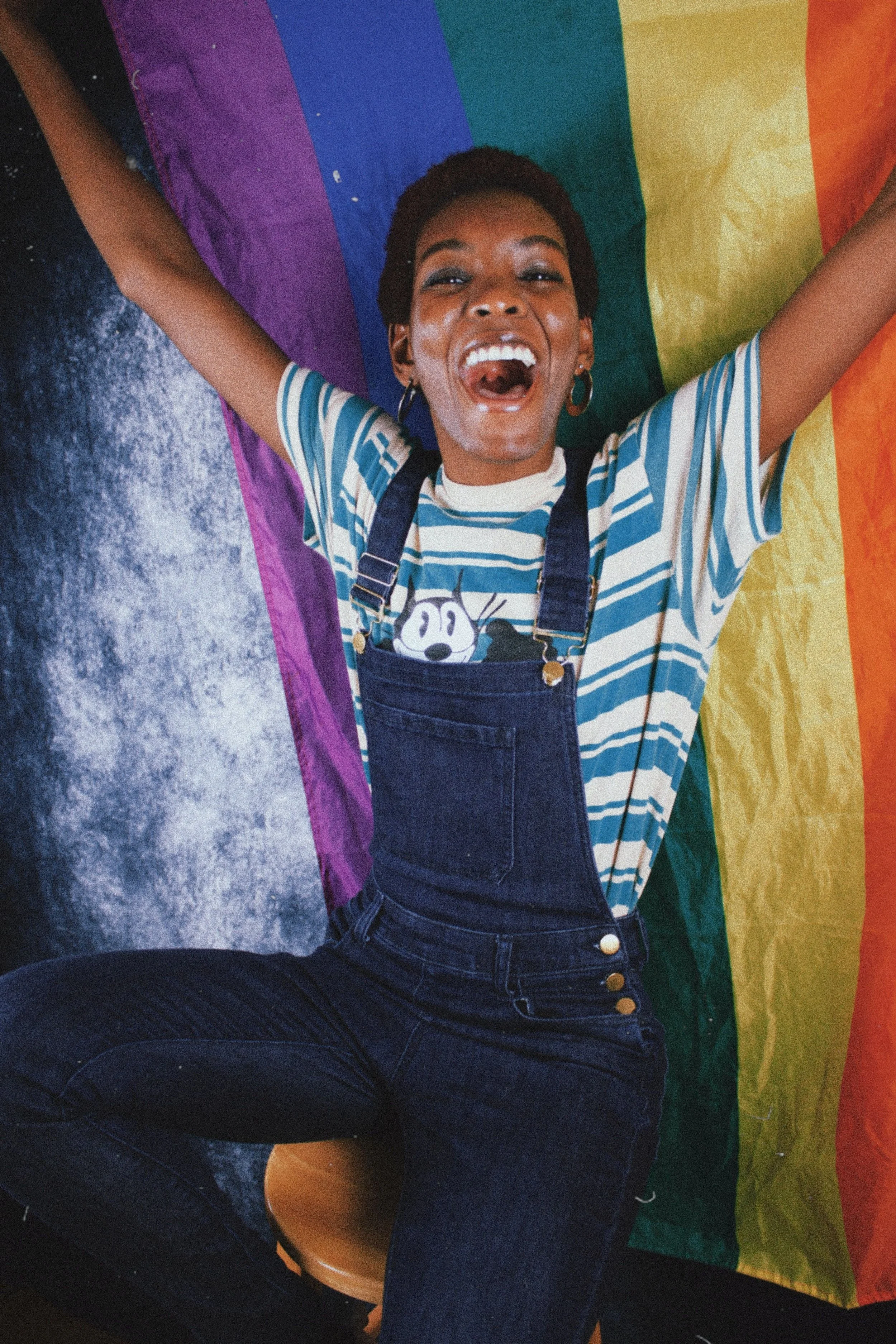 A joyful person with short hair and hoop earrings is celebrating with their arms raised, sitting on a stool against a rainbow pride flag backdrop, wearing a striped T-shirt and denim overalls.