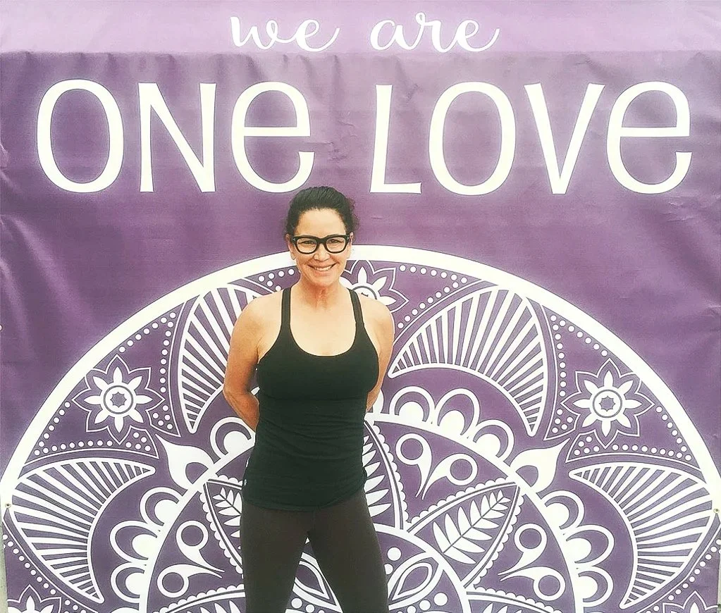 Paula Miller- Austin anxiety therapist standing in front of a purple banner that says 'we are ONE LOVE' with a decorative white mandala design.