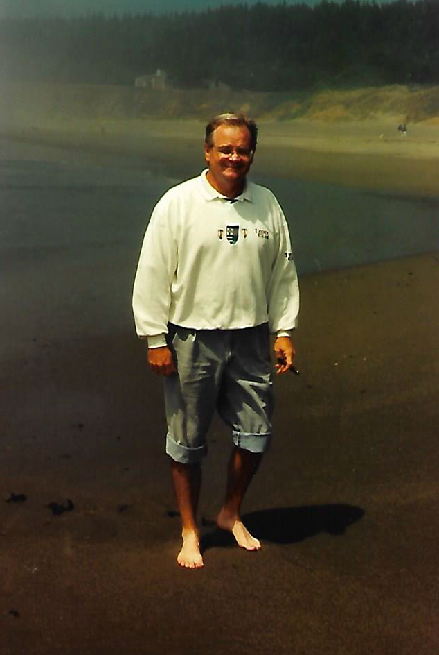 A man standing barefoot on a sandy beach near the water, with a forested hillside in the background, smiling at the camera.